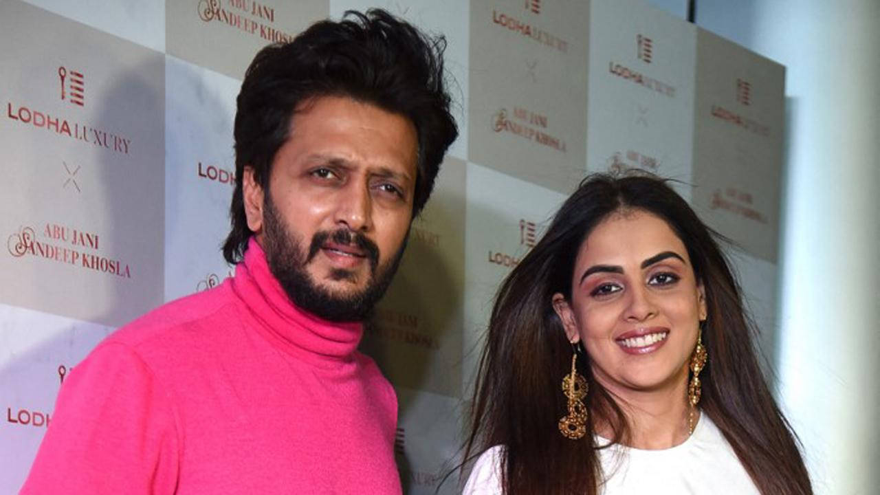 Riteish Deshmukh 與美麗妻子 Genelia 壁紙 — 一名穿著亮粉紅色高領毛衣的男子和一名穿著白色衣服的女子在品牌活動背景前對著鏡頭微笑。
