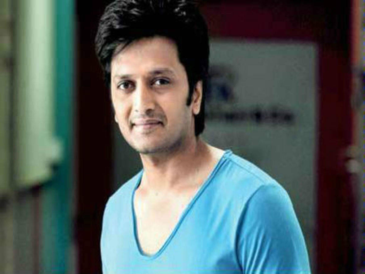 Riteish Deshmukh 簡約造型 壁紙 — 一位深色頭髮、身穿淺藍色V領襯衫的男子，在柔和模糊的室內環境中對著鏡頭溫柔微笑。