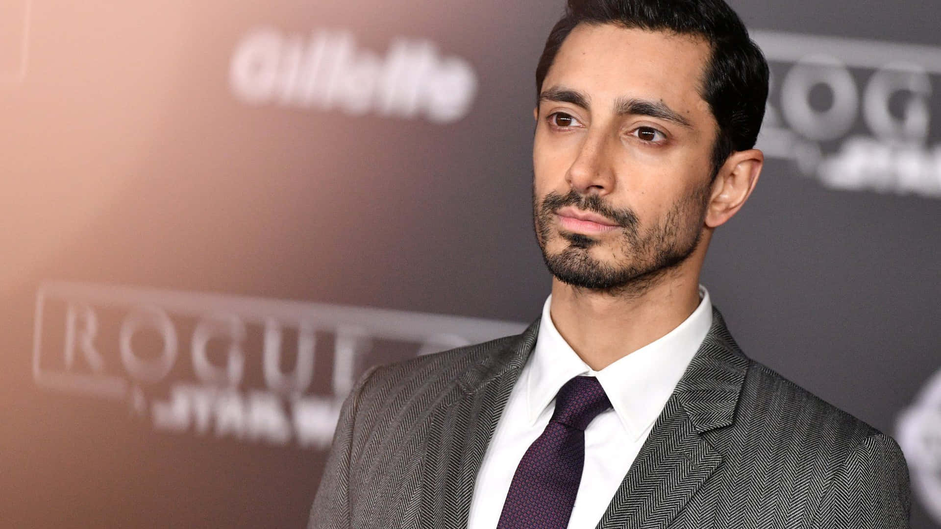Riz Ahmed 活動亮相 壁紙 — 《俠盜一號》演員里茲·阿邁德穿著俐落灰色西裝和紫色領帶，在紅毯活動上，背景為模糊的宣傳佈景。
