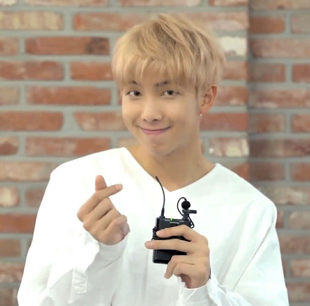 RM BTS 指心手勢 壁紙 — 一位金髮年輕男子穿著白襯衫，微笑著手持黑色無線麥克風，在磚牆背景下做出手指愛心手勢。