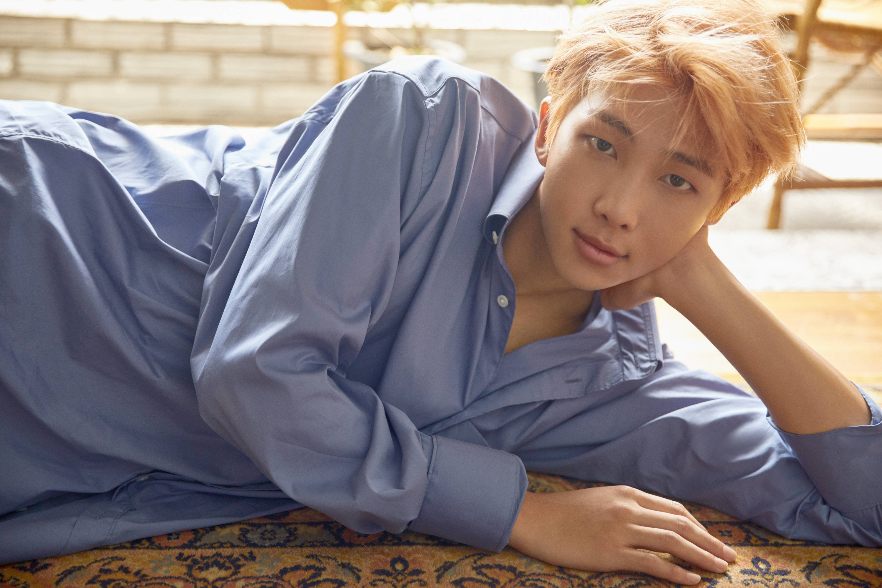 RM BTS《Love Yourself: Her》概念照 壁紙 — 一位金髮年輕男子穿著藍色襯衫，放鬆地躺在圖案地毯上，溫柔地凝視鏡頭。