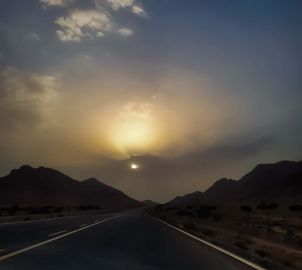 road and sky 壁紙 — 一條長直路在黑暗山脈之間延伸，天空戲劇性，明亮的太陽部分被雲遮擋。