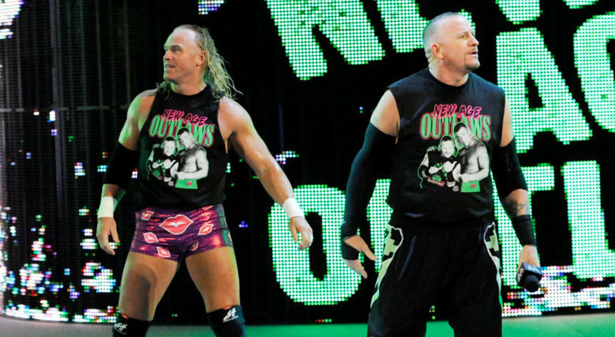 Road Dogg 與比利·岡恩 雙人組合 壁紙 — 兩名肌肉發達的摔角手身穿「New Age Outlaws」裝備，自信地站在燈光明亮的摔角舞台上，身後有發光的綠色LED標牌。