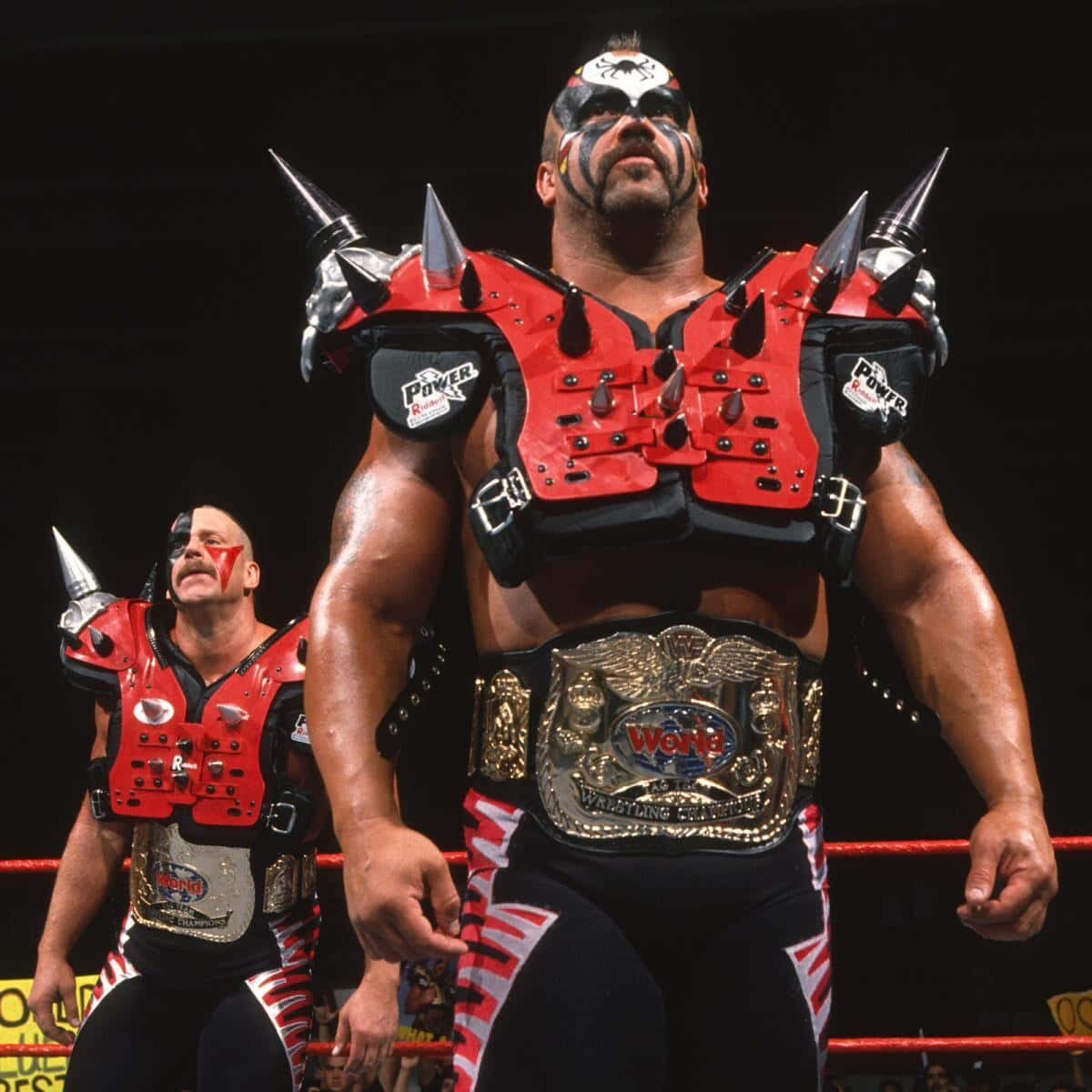 Road Warrior Hawk與Road Warrior Animal奪得WWF雙打冠軍 壁紙 — 兩名肌肉發達的摔角手身穿紅色尖刺盔甲和冠軍腰帶，站在明亮的燈光下的摔角擂台上。