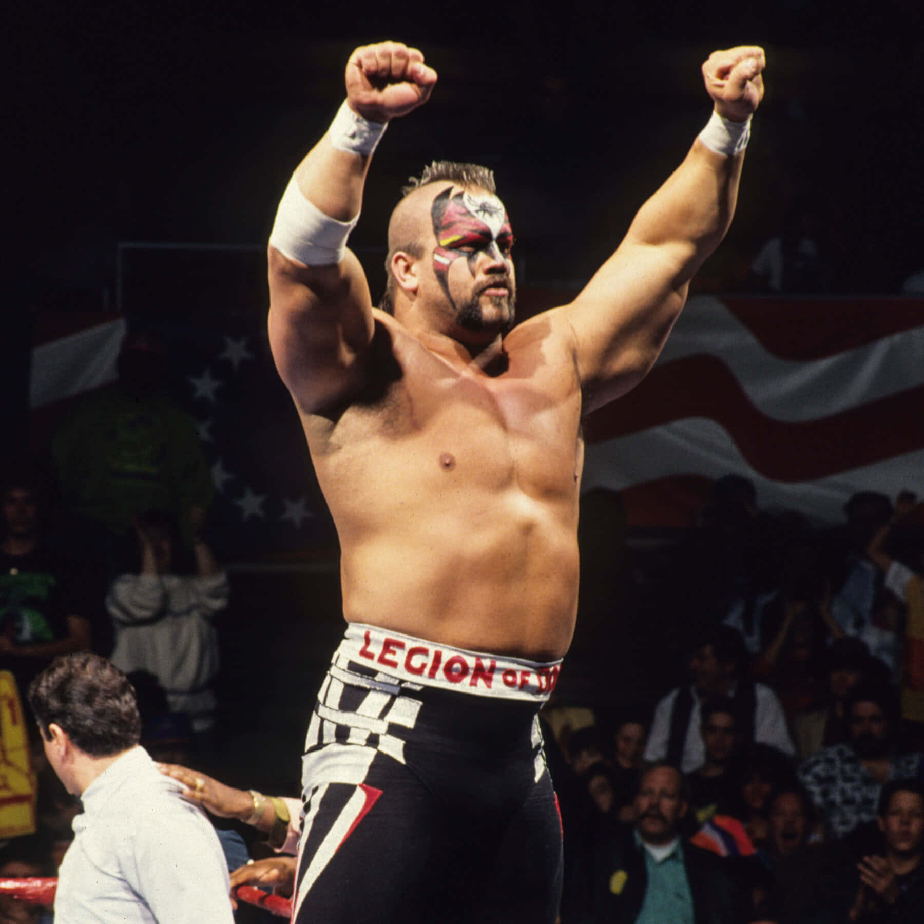 Road Warrior Hawk WWF摔角傳奇 壁紙 — 一位肌肉發達、臉上塗有顏料的摔角手在競技場燈光下高舉雙拳慶祝勝利，身後有群眾和美國國旗。