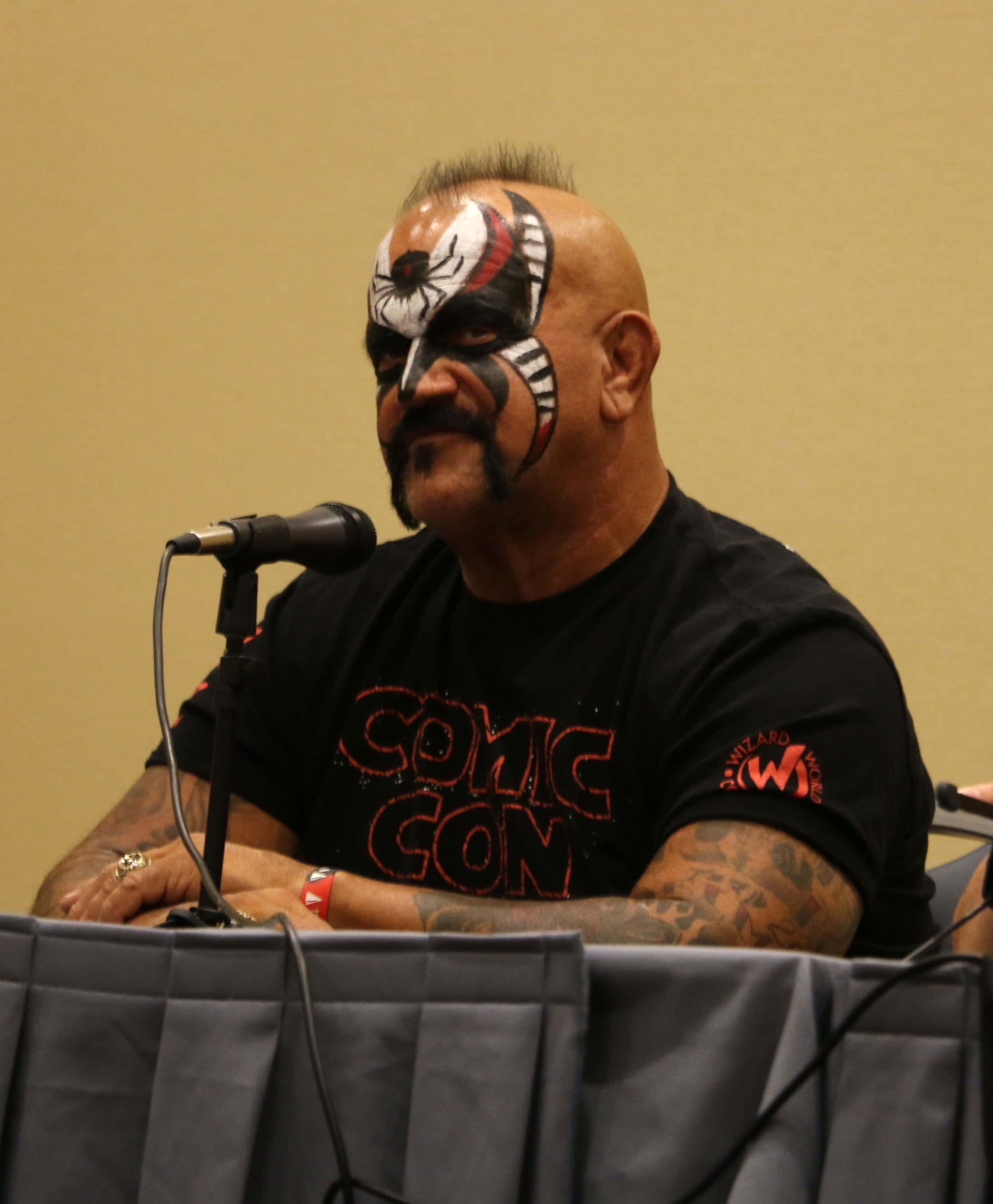 Road Warrior Hawk於芝加哥Wizard World活動 壁紙 — 一名帶有蜘蛛主題面部彩繪的男子坐在漫畫展的桌子旁，面前有一個麥克風。