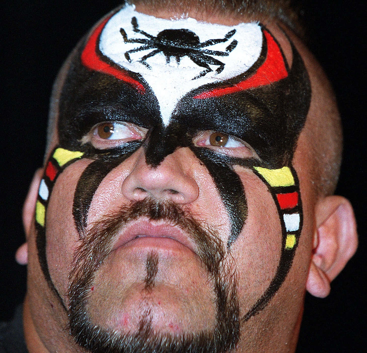 Road Warrior Hawk佩戴標誌性臉部塗裝 壁紙 — 近距離拍攝一名摔角手，臉上塗有醒目的黑色、紅色和黃色顏料，額頭上有一隻黑色蜘蛛。