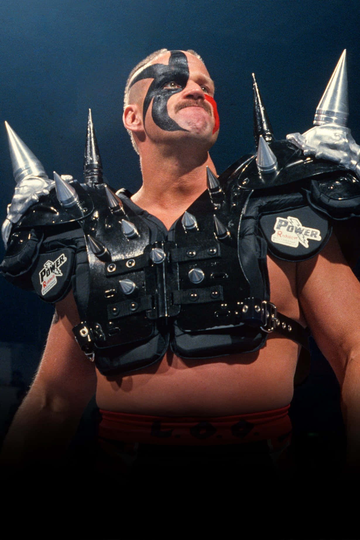 Road Warrior Hawk WWE摔角傳奇 壁紙 — 一名摔角手臉上塗著黑紅色的顏料，身穿帶刺的盔甲，在舞台燈光下自信地微笑向上看。