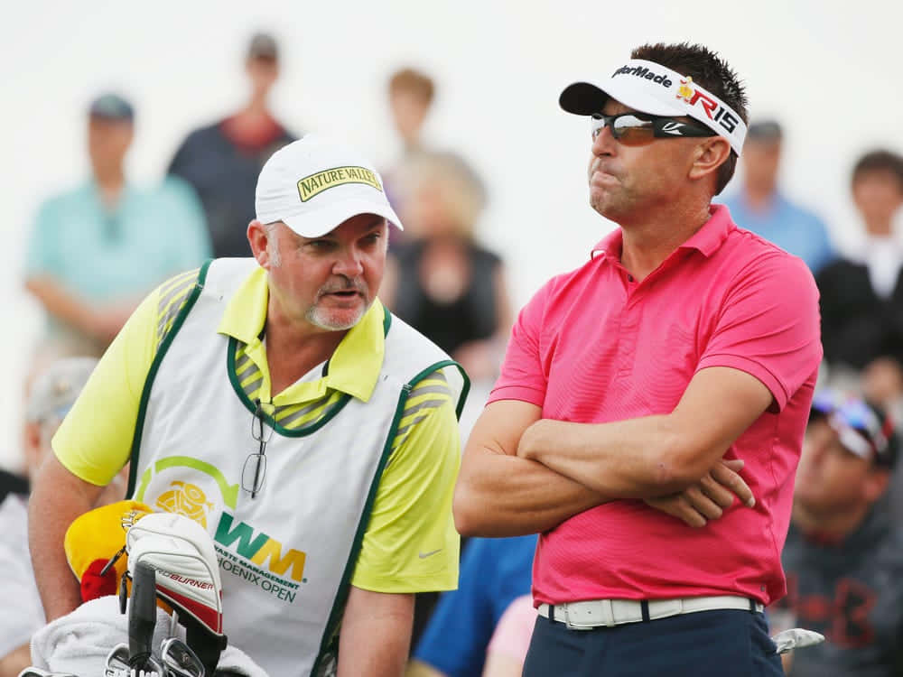 Robert Allenby 身穿粉紅Polo衫 壁紙 — 一位專注的高爾夫球手穿著粉紅色襯衫和遮陽帽，雙手交叉站立，而他的球童穿著黃色襯衫和白色背心，在明亮的球場上在他旁邊說話。