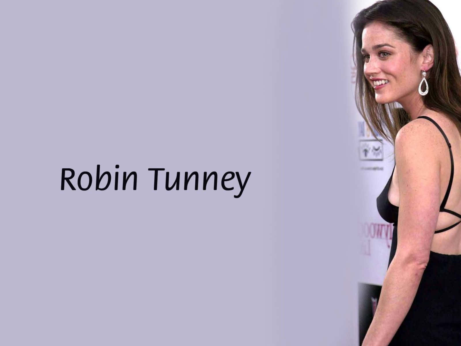 Robin Tunney 桌布 壁紙 — Robin Tunney 身穿黑色連衣裙，在純色薰衣草背景上露出微笑，並顯示她的名字。