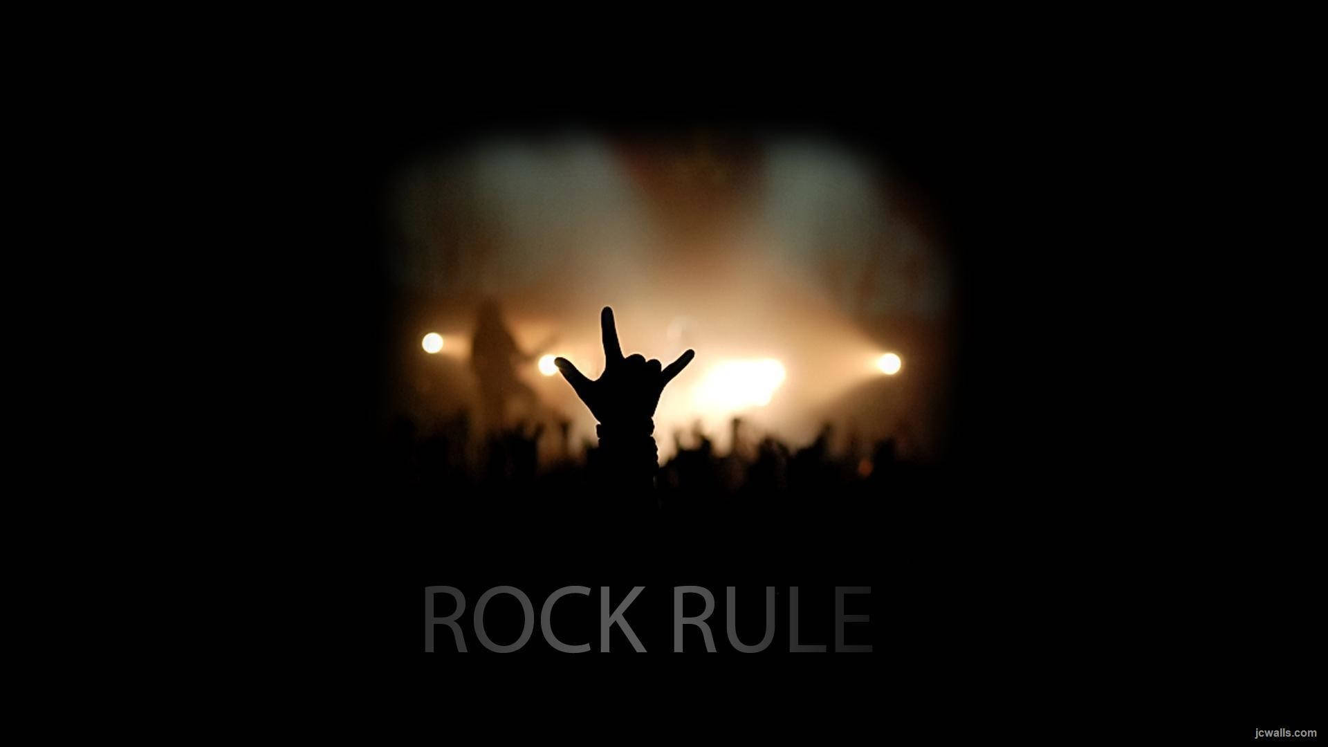 酷炫音樂演唱會中的 Rock 手勢 壁紙 — 一隻剪影手在演唱會中，對著充滿聚光燈和煙霧的發光舞台做出搖滾手勢。