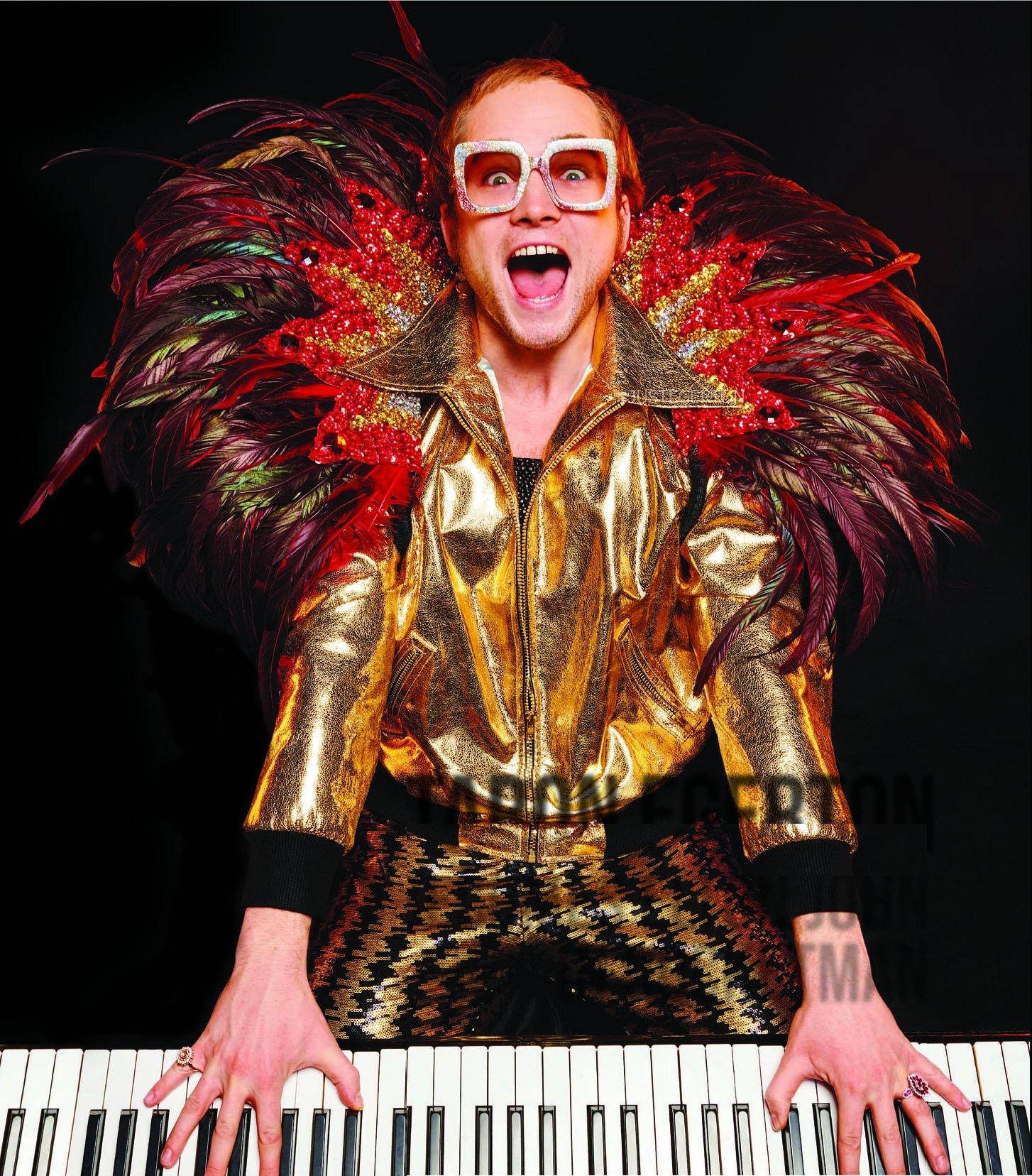 Rocketman 弹奏鋼琴 壁紙 — 一個穿著金色外套和羽毛翅膀的男人在黑色背景上狂喜地彈鋼琴。