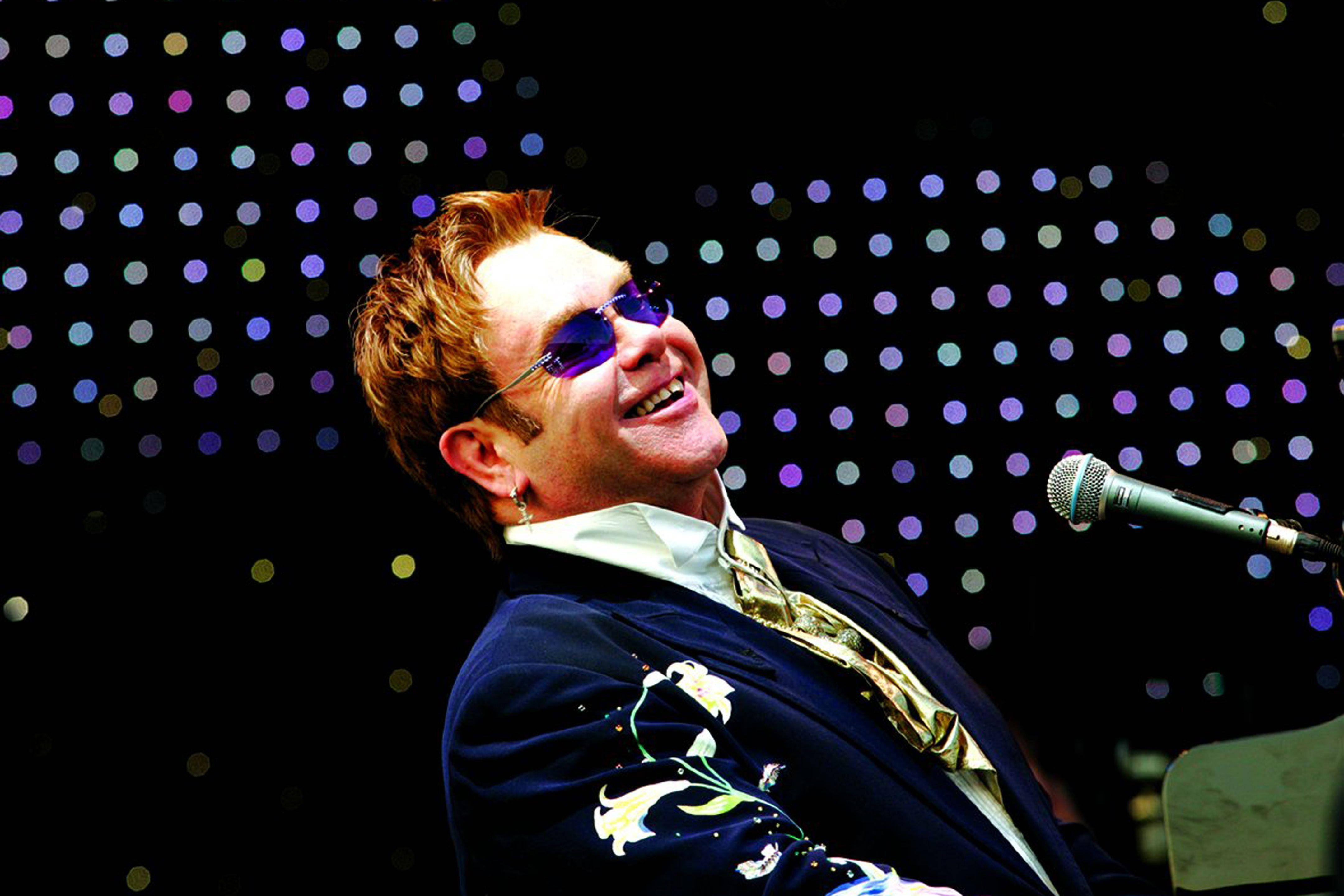 Rocketman 演唱會中開心大笑 壁紙 — 艾爾頓·約翰在舞台上戴著太陽眼鏡和花卉圖案外套，在色彩繽紛的舞台燈光背景下開懷大笑。