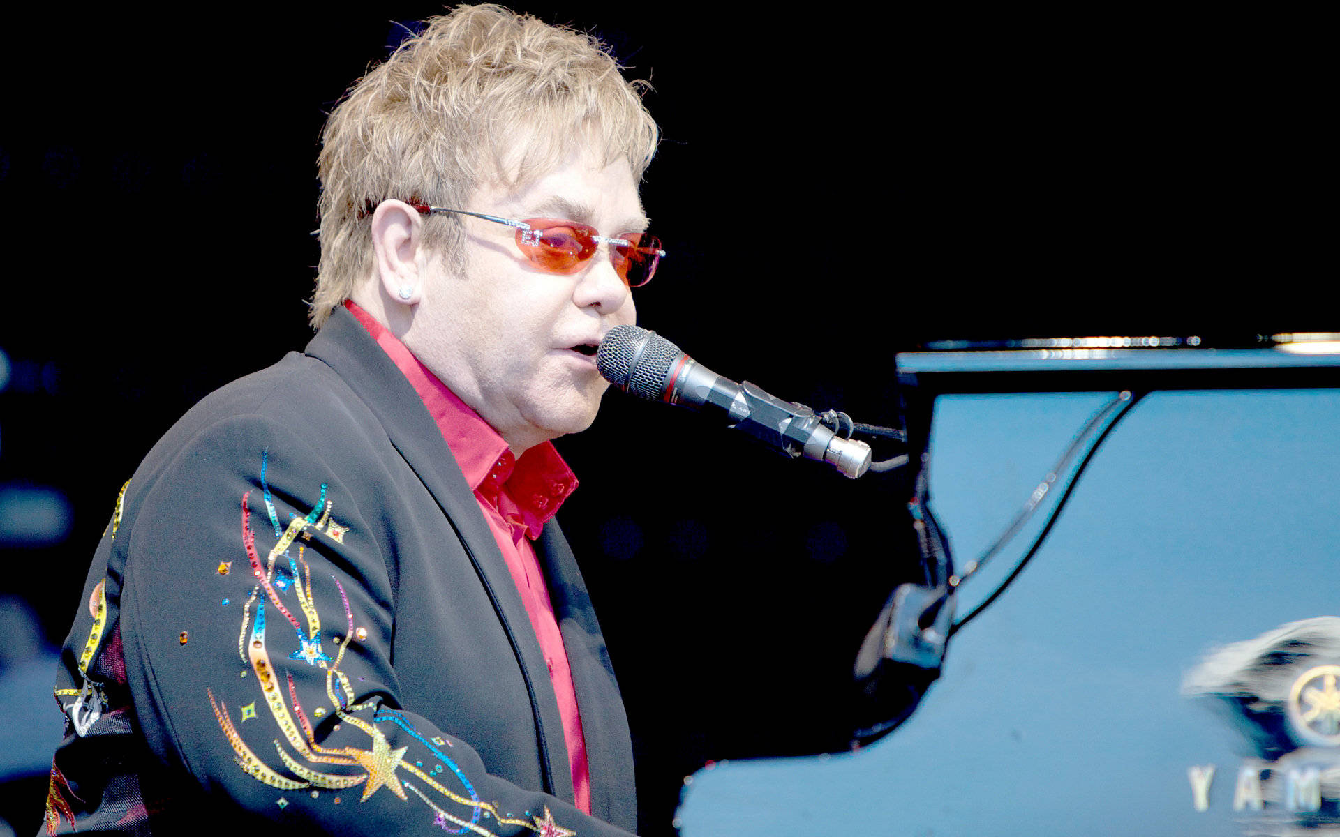 Rocketman 艾爾頓·強演唱 壁紙 — 艾頓莊在舞台燈光下，穿著色彩繽紛的刺繡外套和紅色太陽眼鏡，熱情地彈奏鋼琴。