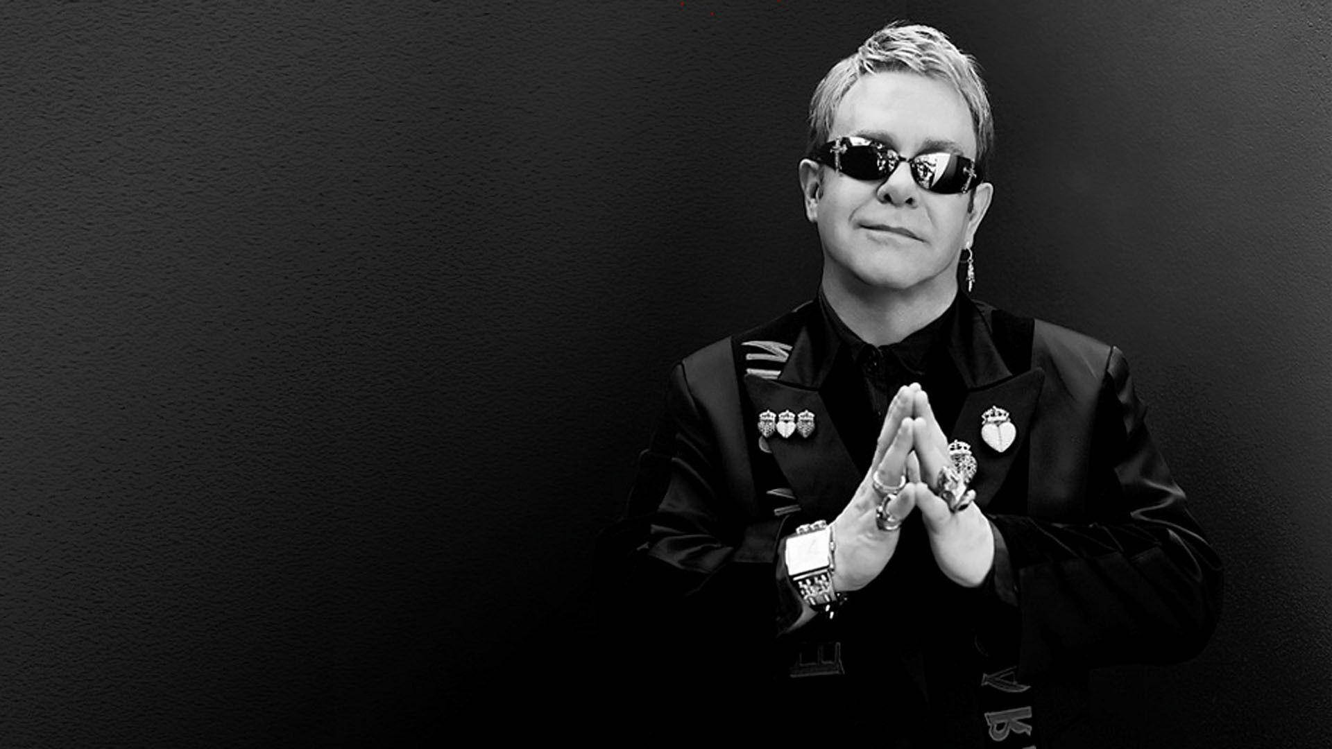 Rocketman 黑白肖像 壁紙 — 艾頓莊穿著黑色西裝、戴著太陽眼鏡，雙手合攏，在深色紋理背景前擺姿勢。