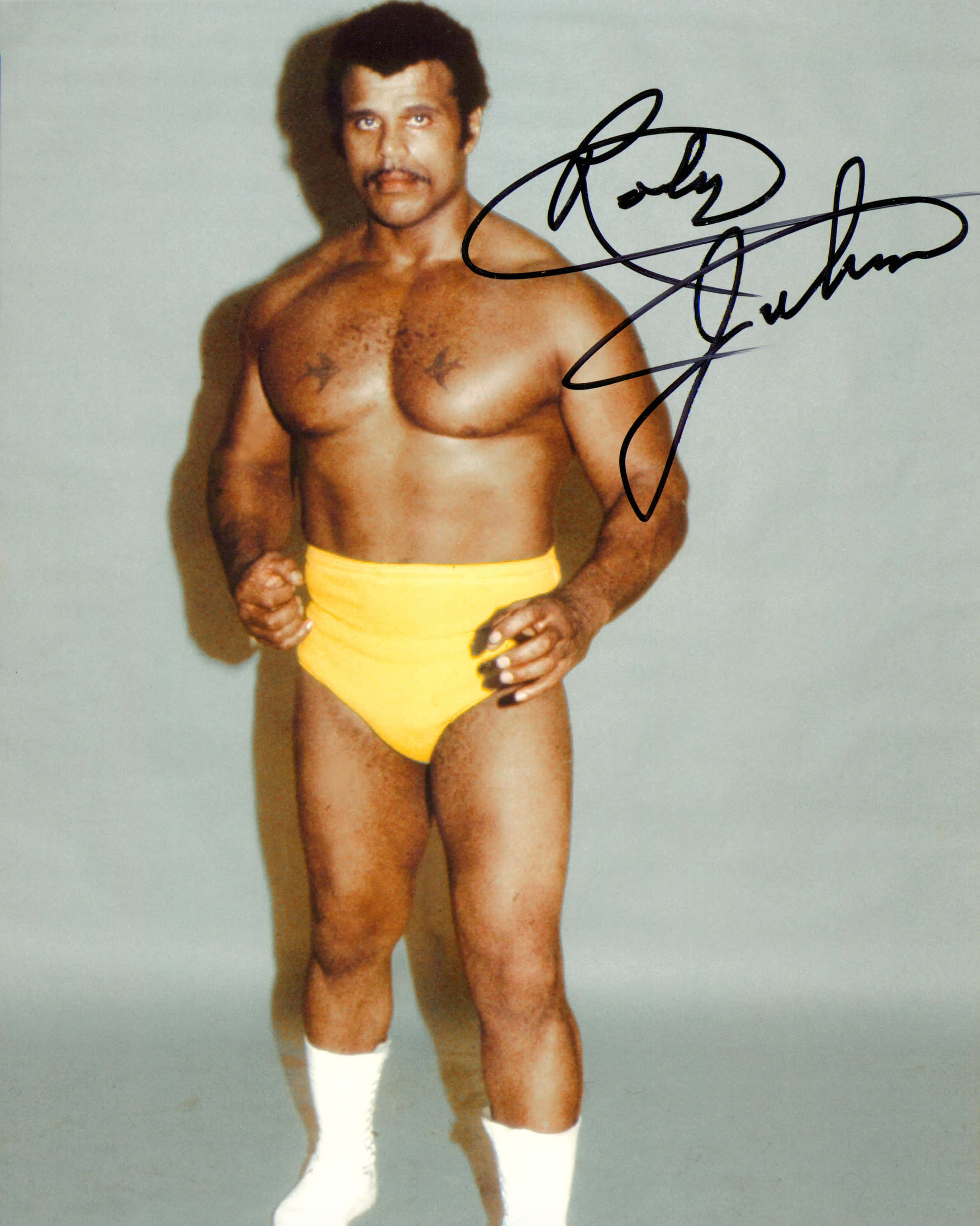 Rocky Johnson 簽名照 壁紙 — 肌肉發達的摔角手 Roly Green 身穿黃色短褲和白色襪子，在純色攝影棚背景前自信地擺出姿勢，並附有簽名。