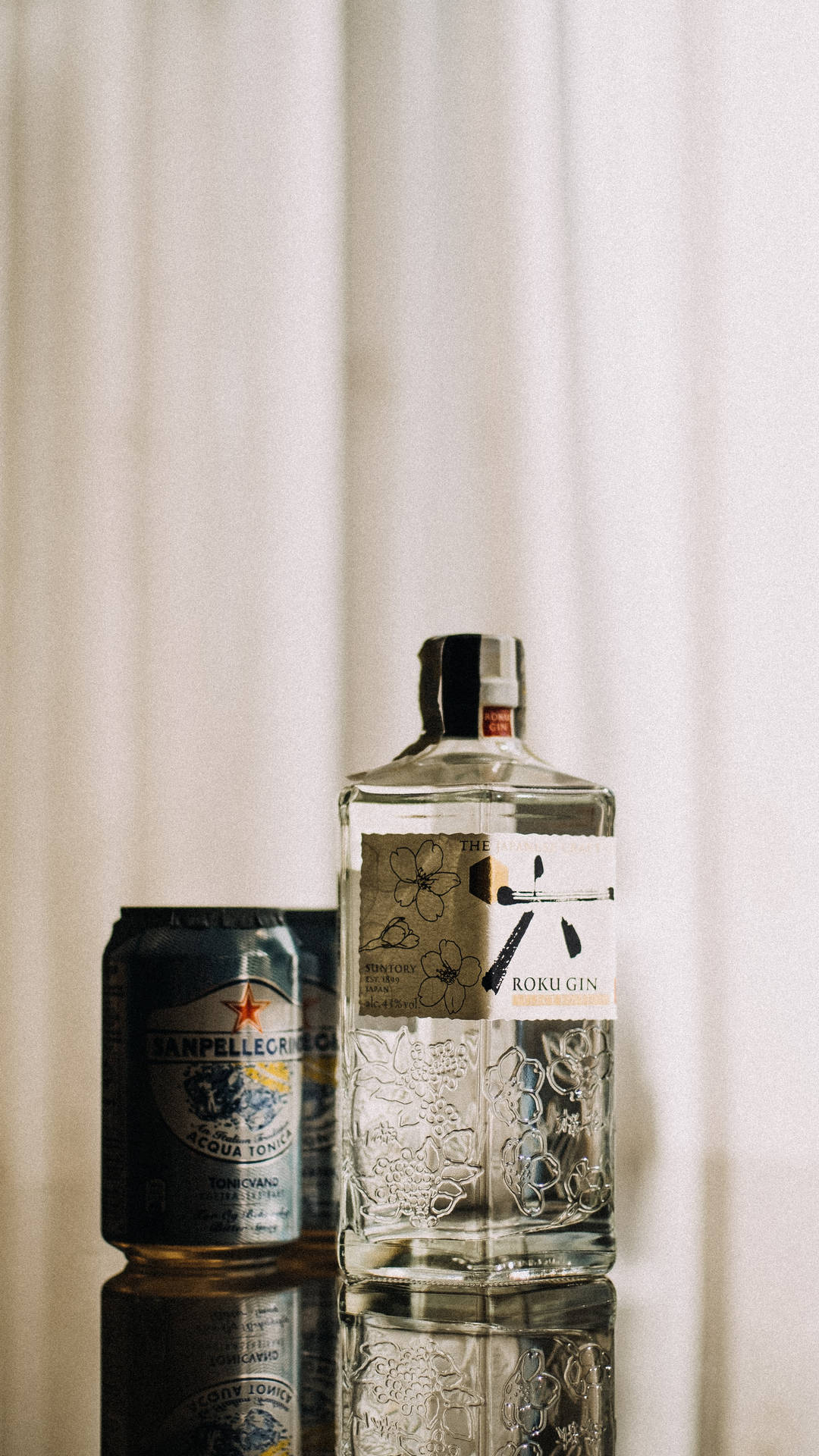 一瓶Roku Gin和San Pellegrino tonic放在反射表面上，後面有白色窗簾。