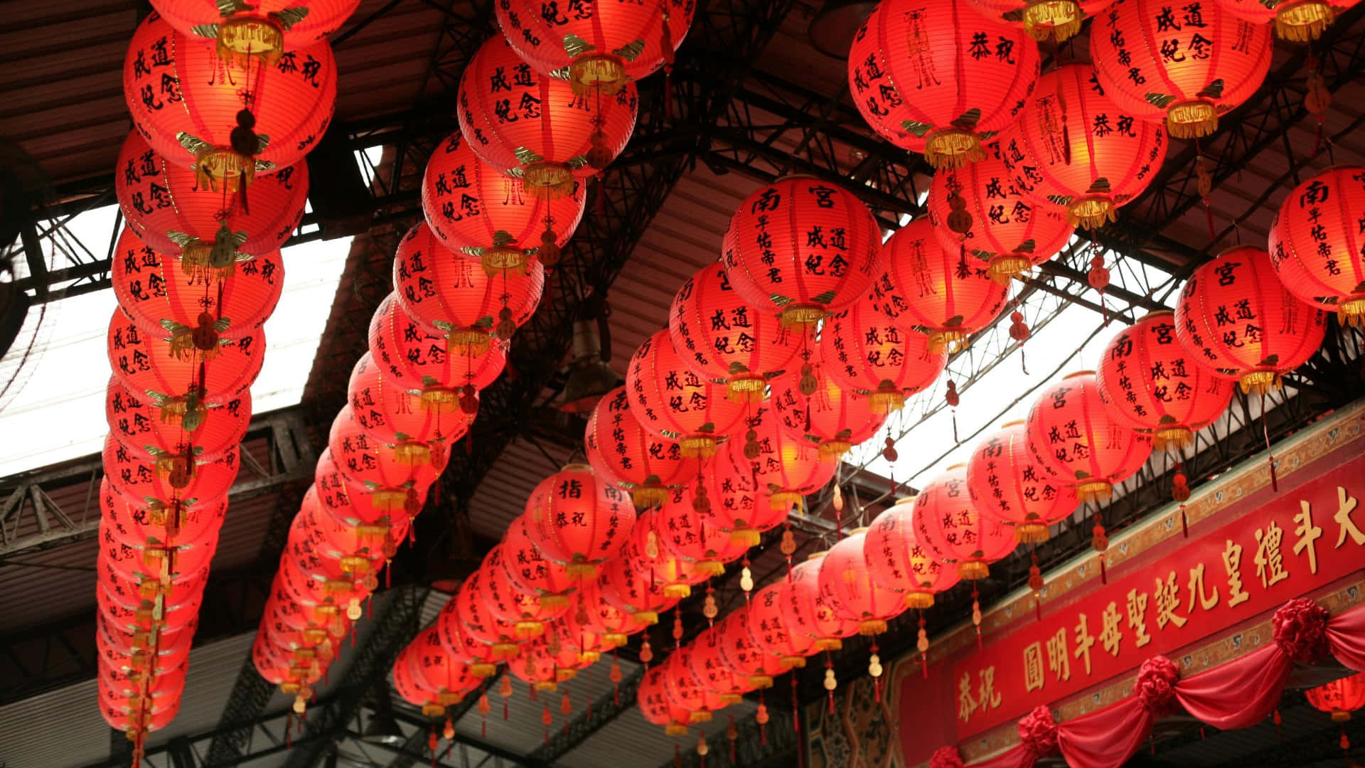  壁紙 — 一排排發光的紅色中國燈籠，帶有黑色書法，密密麻麻地懸掛在金屬屋頂結構上。