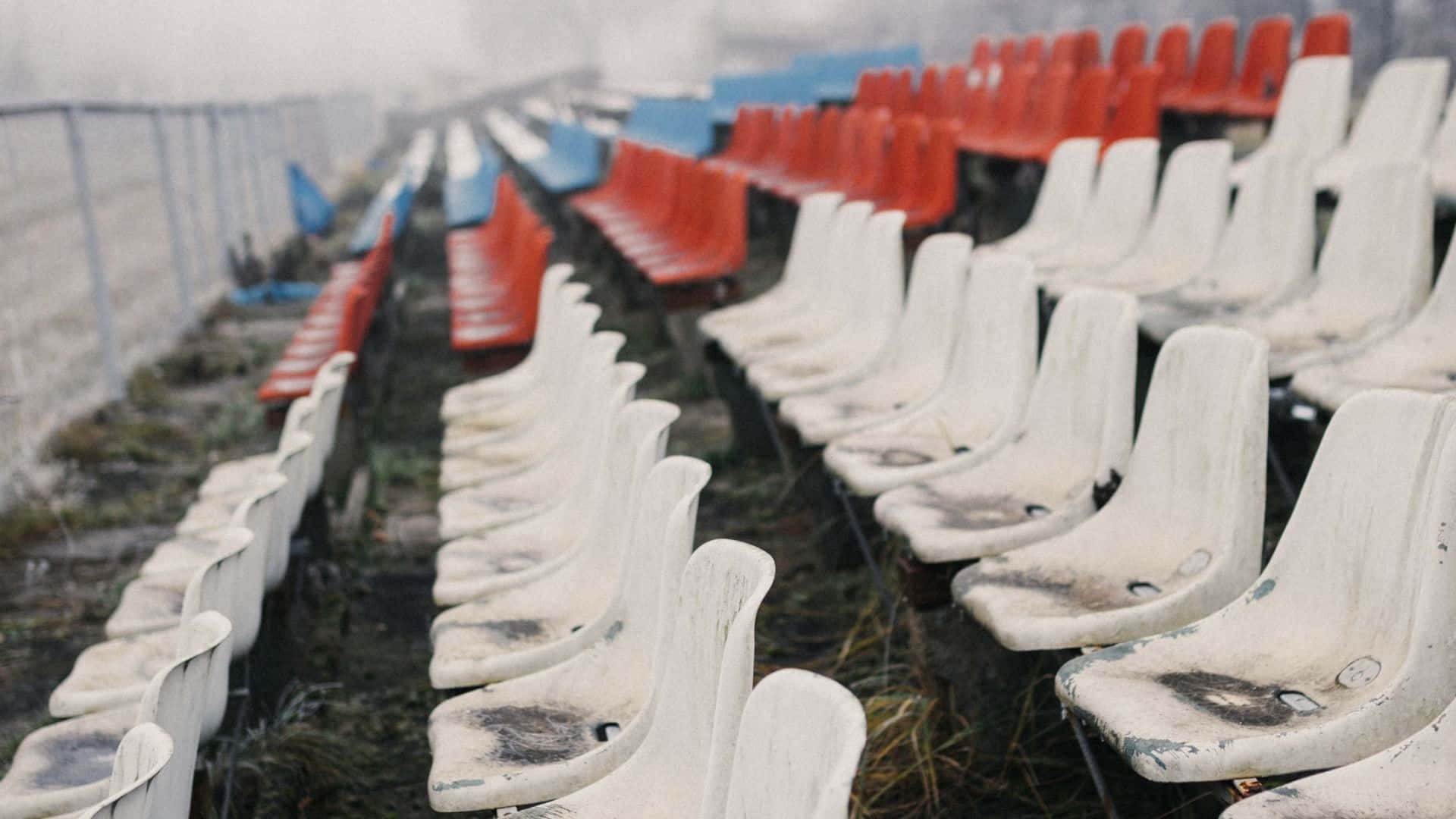  壁紙 — 一排排經風化的白色、紅色和藍色體育場座位，被遺棄在雜草叢生的草地上，天空朦朧。