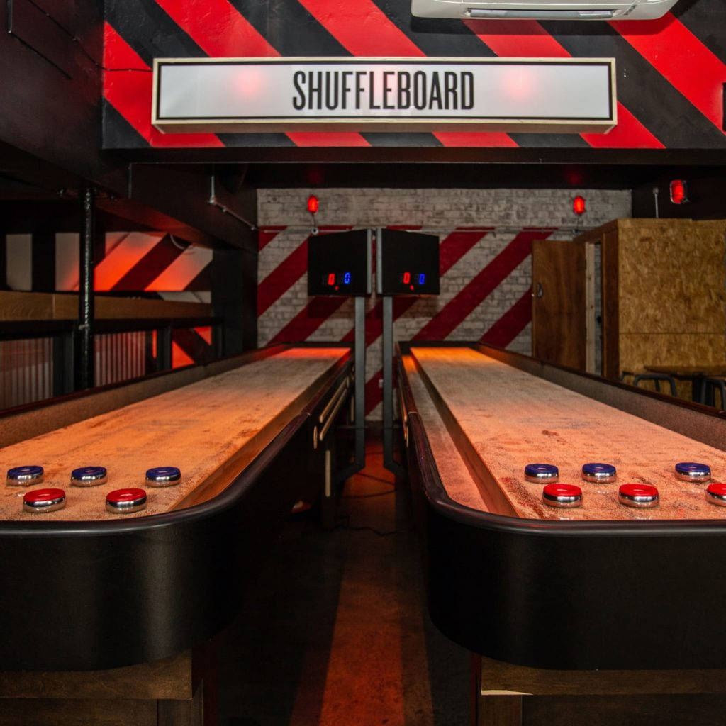 Roxy Ball Room 萊斯特梅里昂街 Shuffleboard 壁紙 — 兩張沙狐球桌，上面有紅色和藍色的冰球，在溫暖的燈光下，位於有條紋牆壁的復古遊戲廳。
