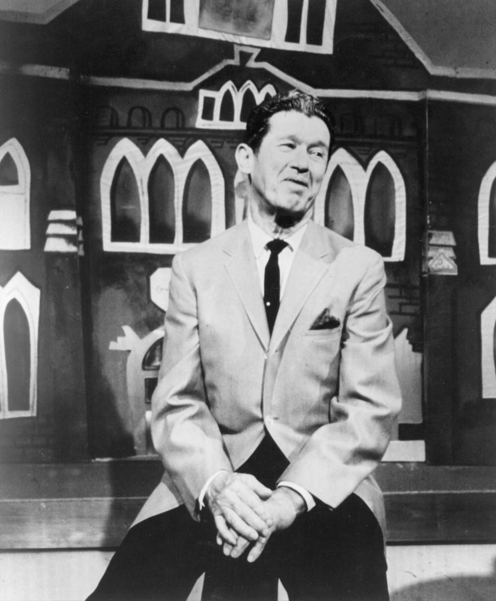 Roy Acuff 唱歌與演出 壁紙 — 一名身穿西裝的男子在復古黑白照片中，平靜地坐在風格化的卡通屋背景前。