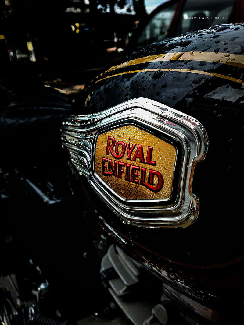 標準 Royal Enfield 標誌 壁紙 — 近距離拍攝濕漉漉的Royal Enfield電單車油箱，鍍鉻徽章在昏暗燈光下反射雨滴。