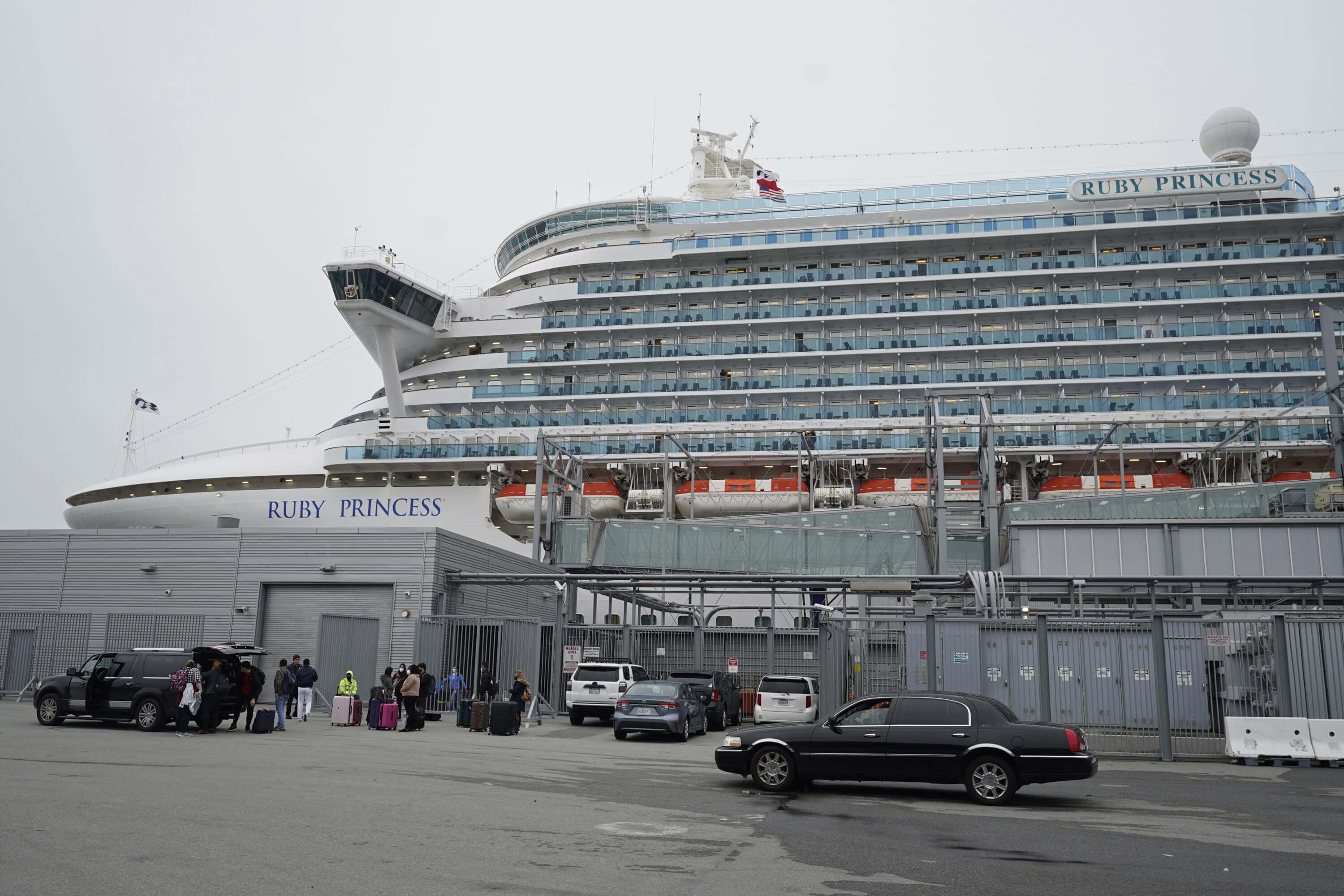 Cruise Ship 桌布 壁紙 — Ruby Princess 郵輪在港口顯得龐大，乘客和汽車聚集在灰色客運大樓附近，天空多雲。