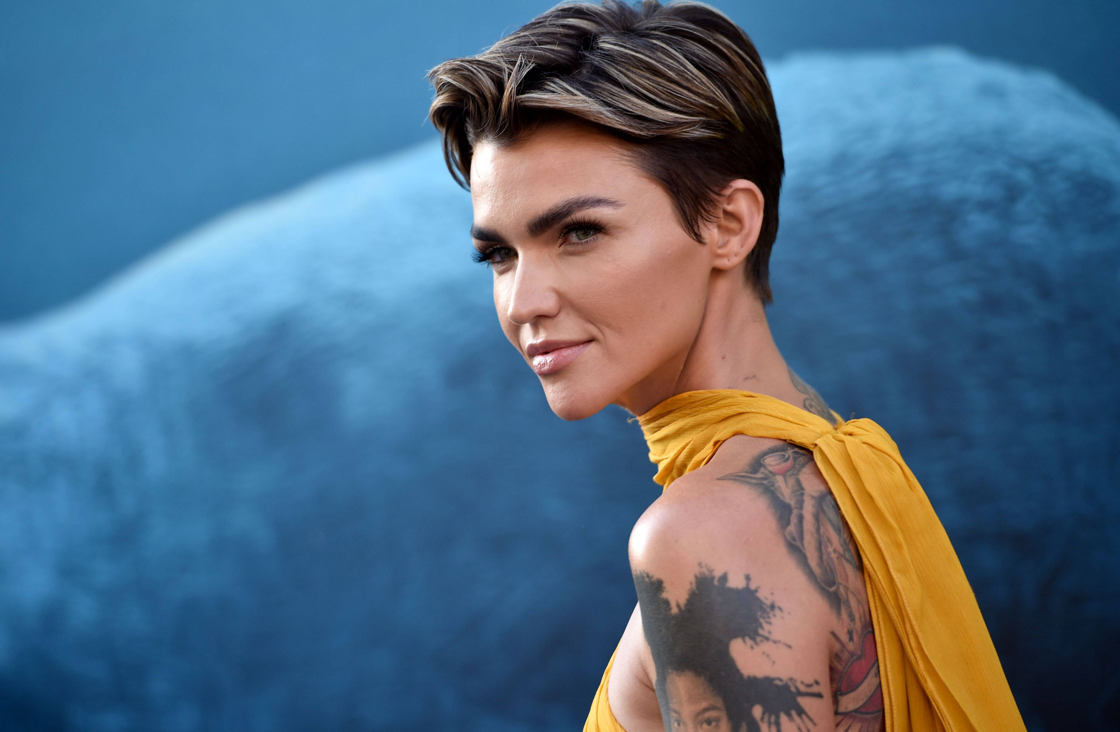 Ruby Rose《巨齒鯊》首映禮 壁紙 — 一位短髮造型、有明顯紋身的女性，穿著黃色垂墜上衣，背景為有紋理的藍色。