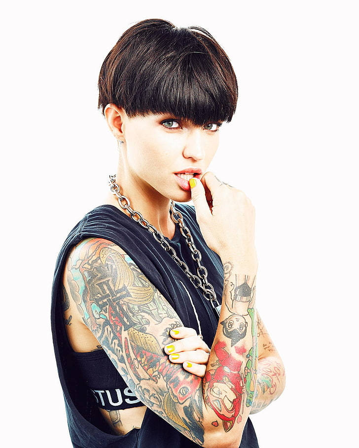 Ruby Rose 碗髮造型 壁紙 — 一位有紋身、短黑髮、妝容大膽的女性，咬著塗黃色的指甲，戴著鏈條項鍊和無袖上衣。