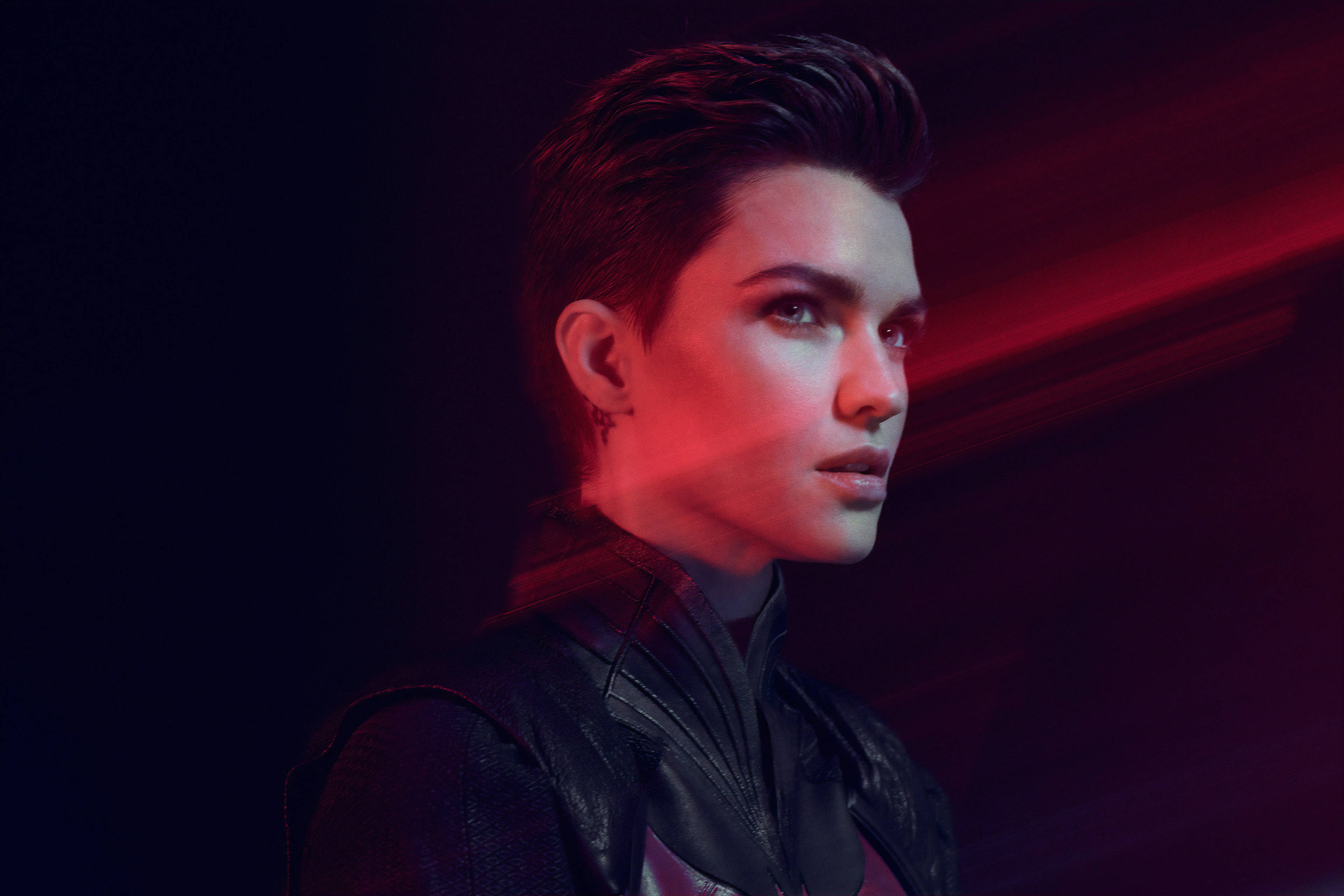 Ruby Rose 展現獨特蓬鬆髮型 壁紙 — 一個短髮、身穿皮褸嘅人喺黑暗背景前，喺戲劇性嘅紅色燈光下向前凝視。