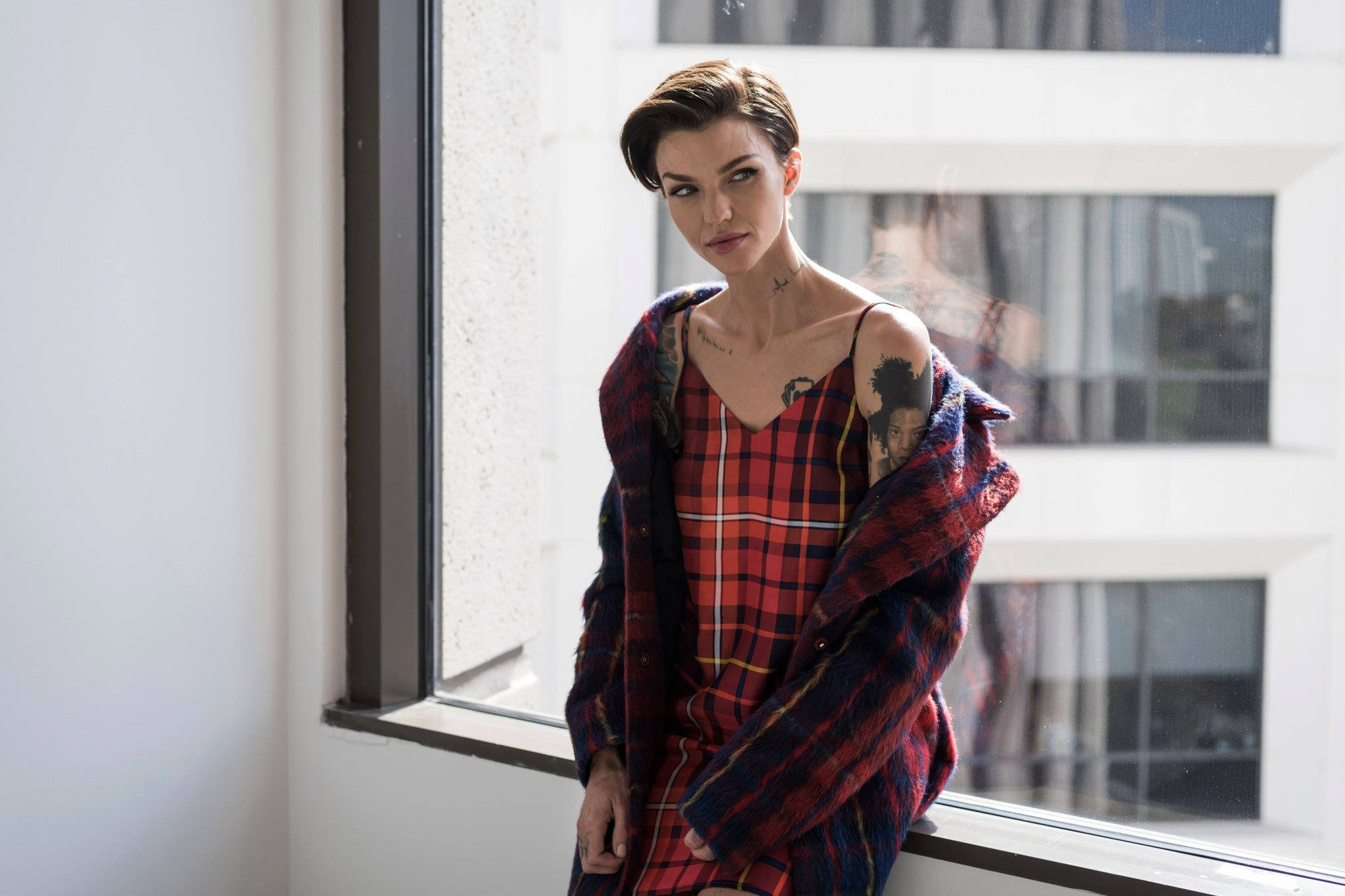Ruby Rose 穿休閒連衣裙 壁紙 — 一位短髮且有紋身的女士坐在窗邊，身穿紅色格子連衣裙和相配的夾克。