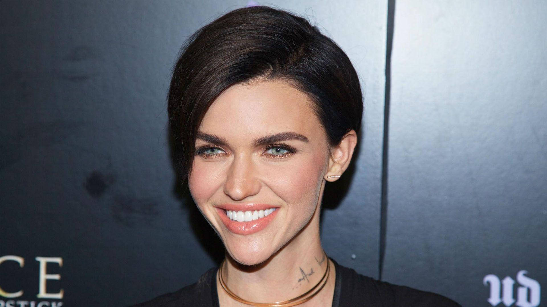 Ruby Rose 經典 pixie 剪髮 壁紙 — 一位短黑髮、明亮藍眼睛的女性在帶有標誌的深色背景下溫暖地微笑。