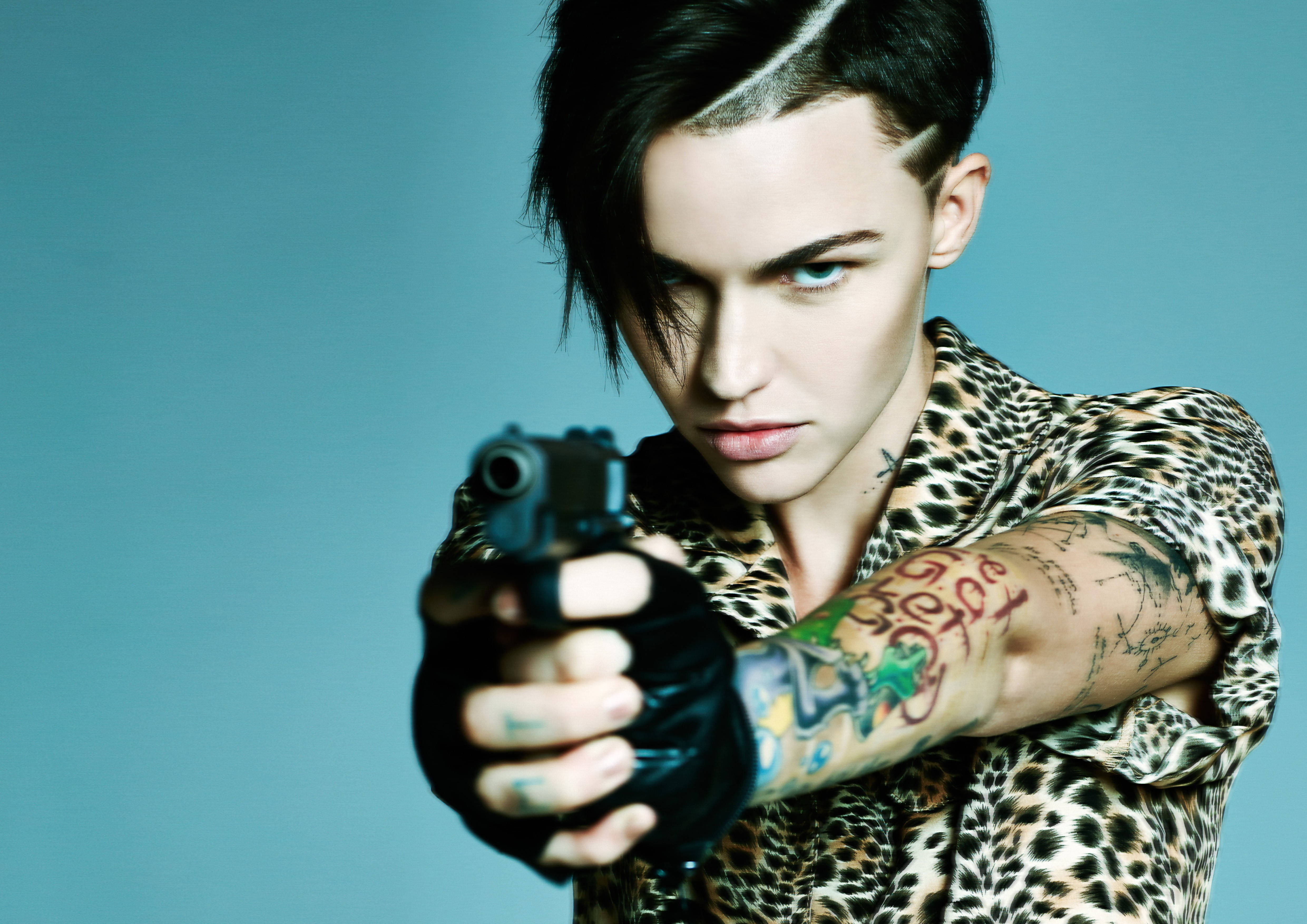 Ruby Rose 飾《生化危機》阿比蓋爾 壁紙 — 一個有紋身、穿著豹紋襯衫的人，在純藍色背景下直接將槍指向觀眾。