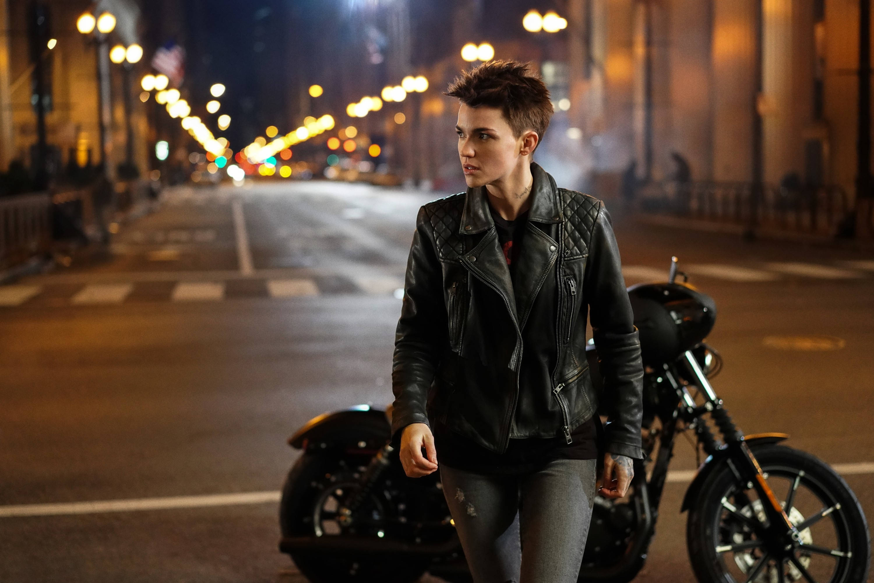 Ruby Rose 飾演 Batwoman Kate Kane 壁紙 — 一個穿著黑色皮褸的人喺夜晚行喺一架電單車旁邊，喺一條濕漉漉嘅城市街道上，街燈閃爍。