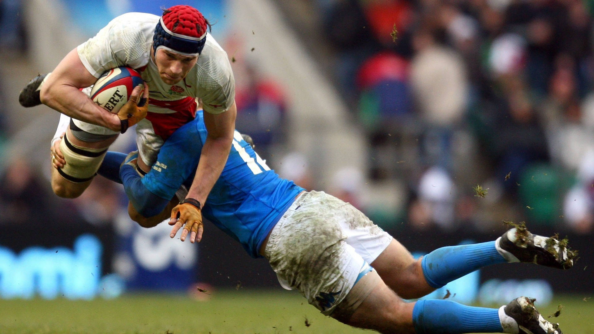 英格蘭rugby 擊球瞬間 壁紙 — 一名身穿紅白色球衣的欖球球員在泥濘的場地上被身穿藍色球衣的對手在半空中攔截時緊緊抓住球。