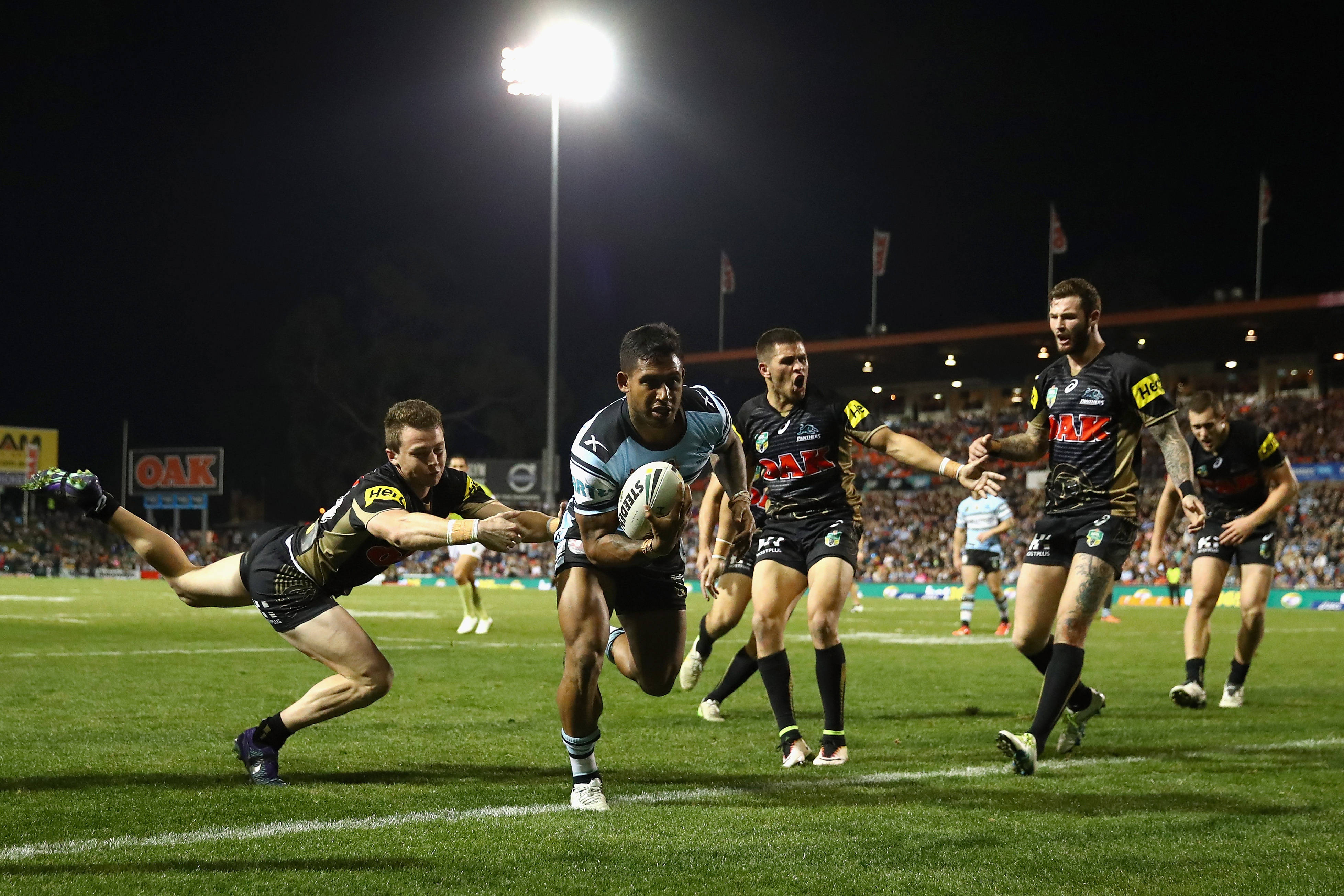 超級聯賽本·巴爾巴 rugby 壁紙 — 一名身穿淺藍色球衣的欖球聯賽球員在夜間明亮的球場燈光下帶球衝刺，防守球員正在攔截他。