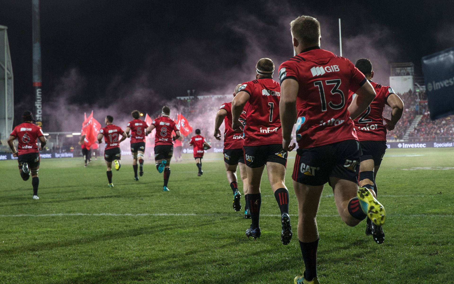 克魯薩德斯 rugby 隊 壁紙 — 身穿紅色球衣的欖球運動員在夜間衝過綠色球場，身後煙霧和紅旗飄揚。