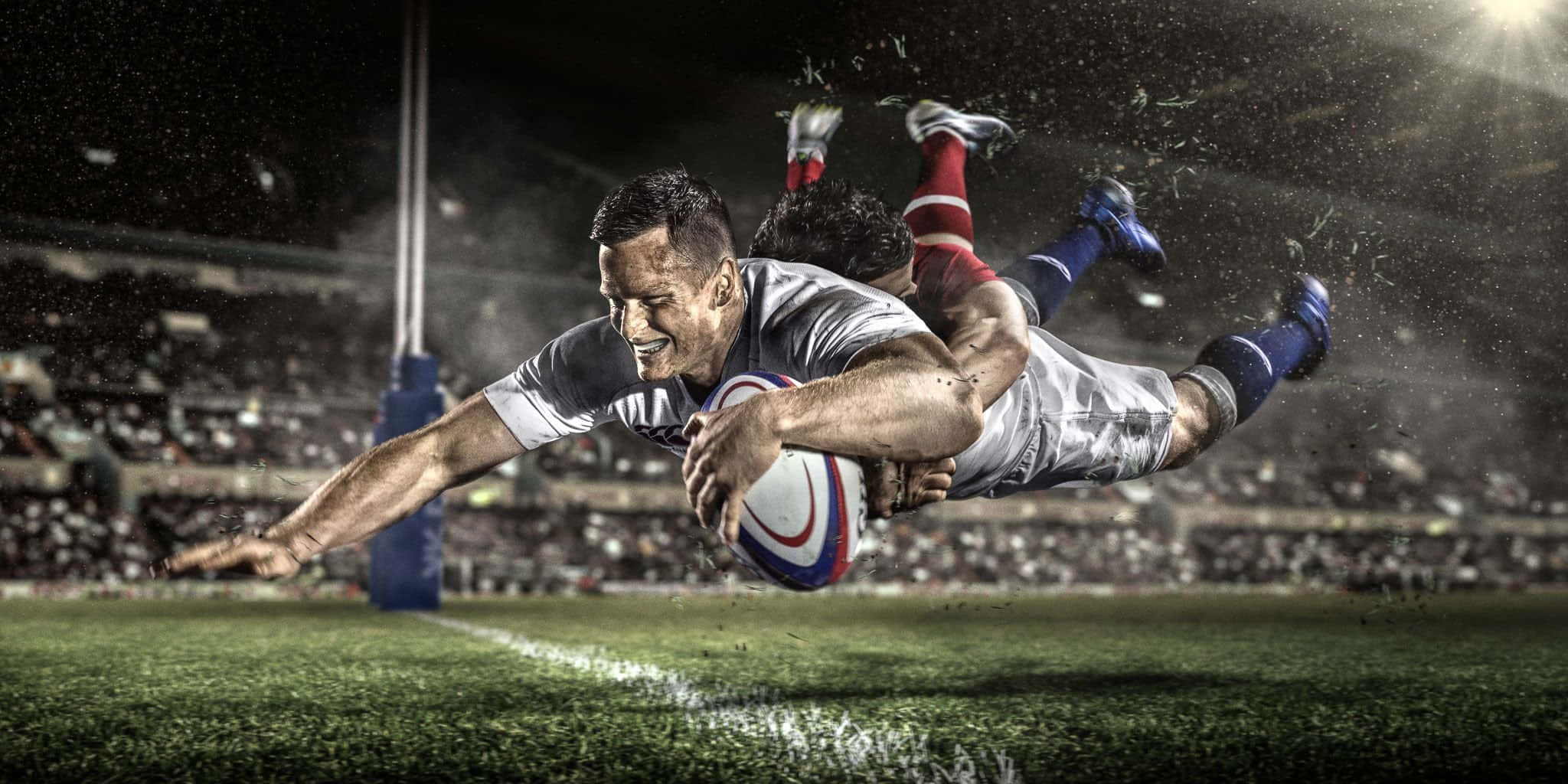 Rugby 桌布 壁紙 — 一名欖球運動員飛身撲向達陣線，防守球員正在攔截他，這是一個戲劇性的夜間球場場景。