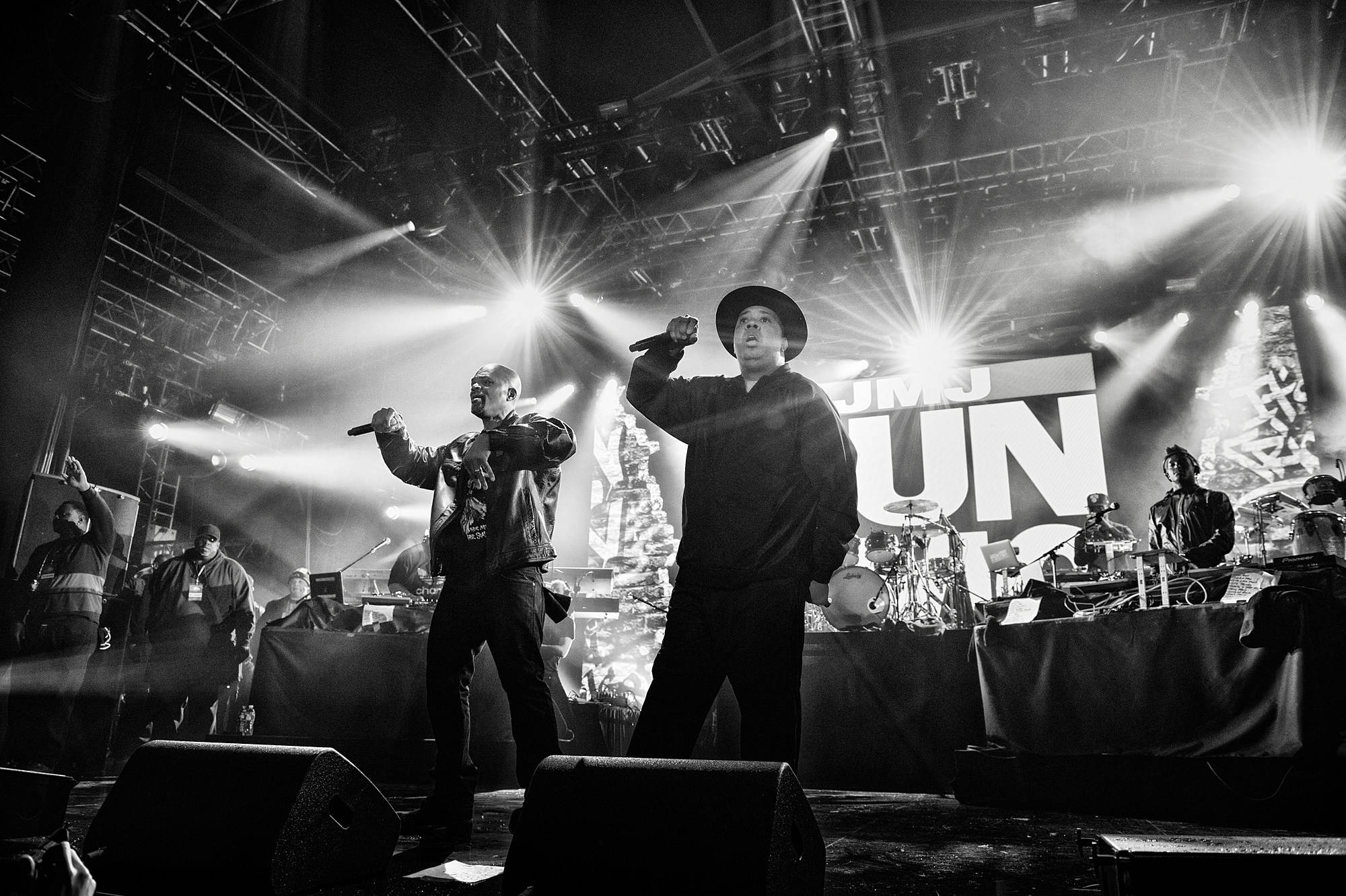 Run D.M.C. 跳躍演唱會 壁紙 — 兩位說唱歌手在燈光昏暗的舞台上，沐浴在戲劇性的聚光燈下，身後有樂隊，充滿活力地表演。