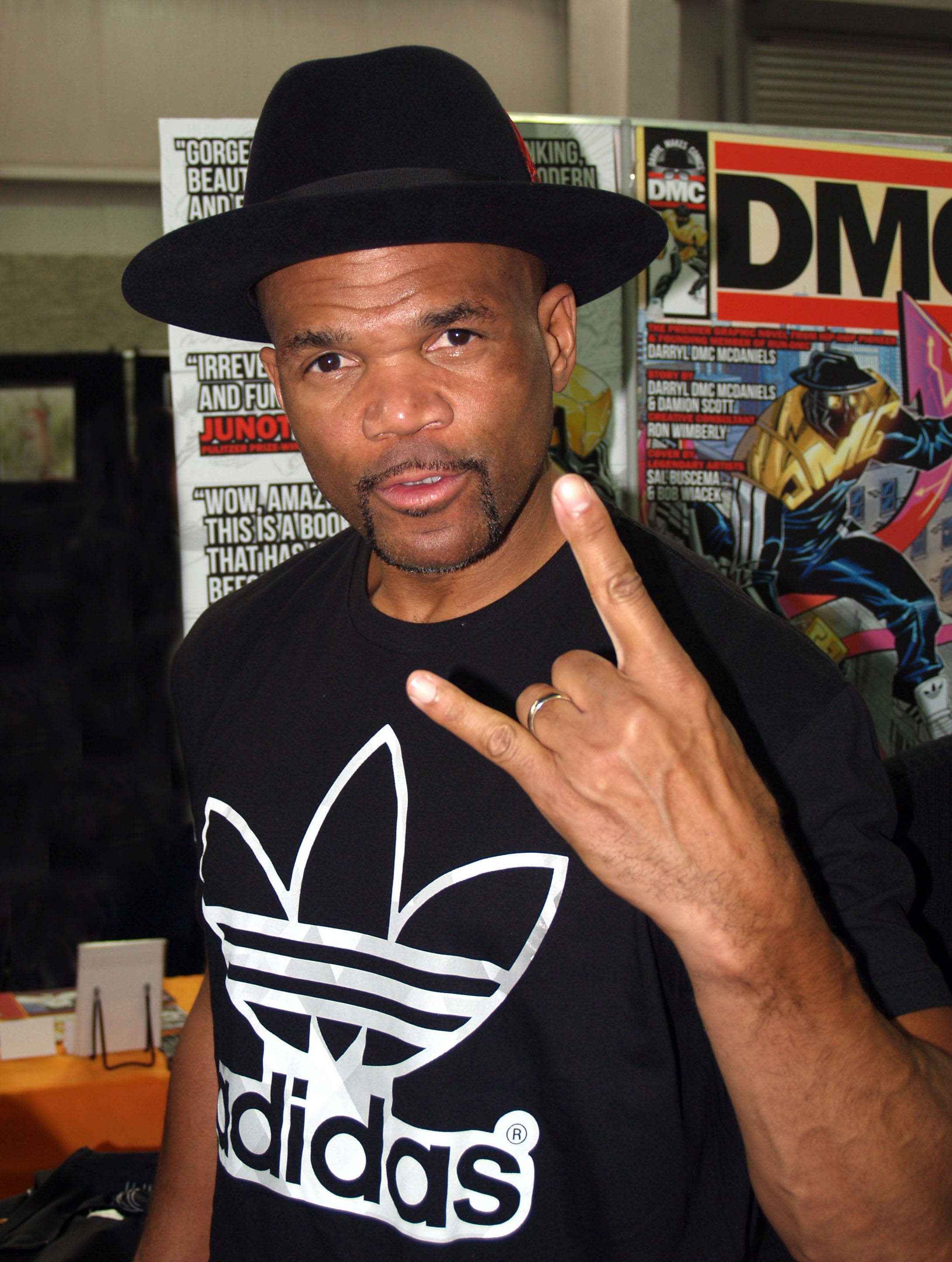 Run D.M.C. 嘻哈藝人 Darryl McDaniels 壁紙 — 一個戴黑色紳士帽、穿Adidas T恤的男人在漫畫海報前做出搖滾手勢。