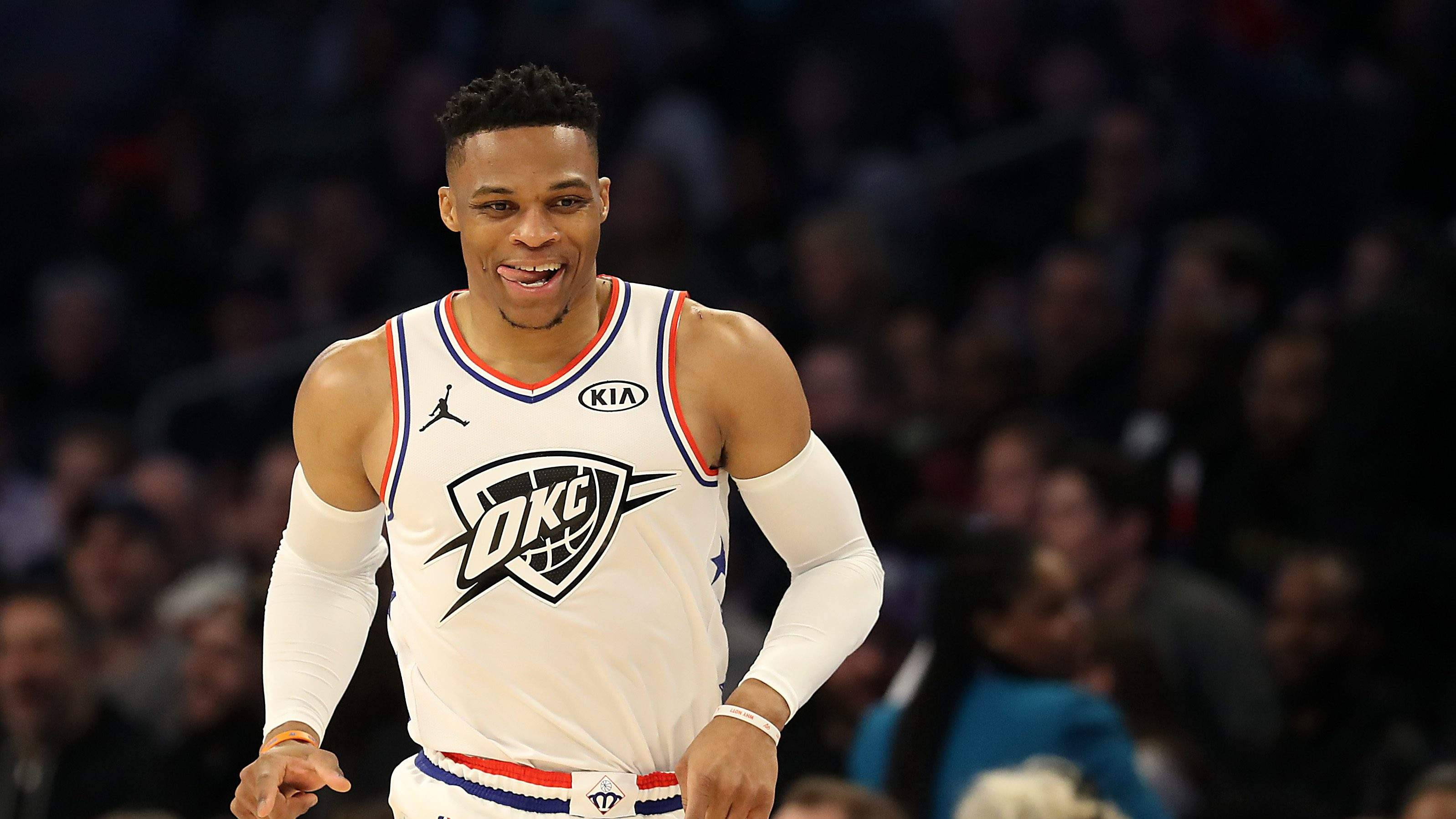 Russell Westbrook 吐舌頭奔跑 壁紙 — 一名身穿白色OKC球衣的籃球運動員在比賽中咧嘴大笑，身後是模糊的人群。