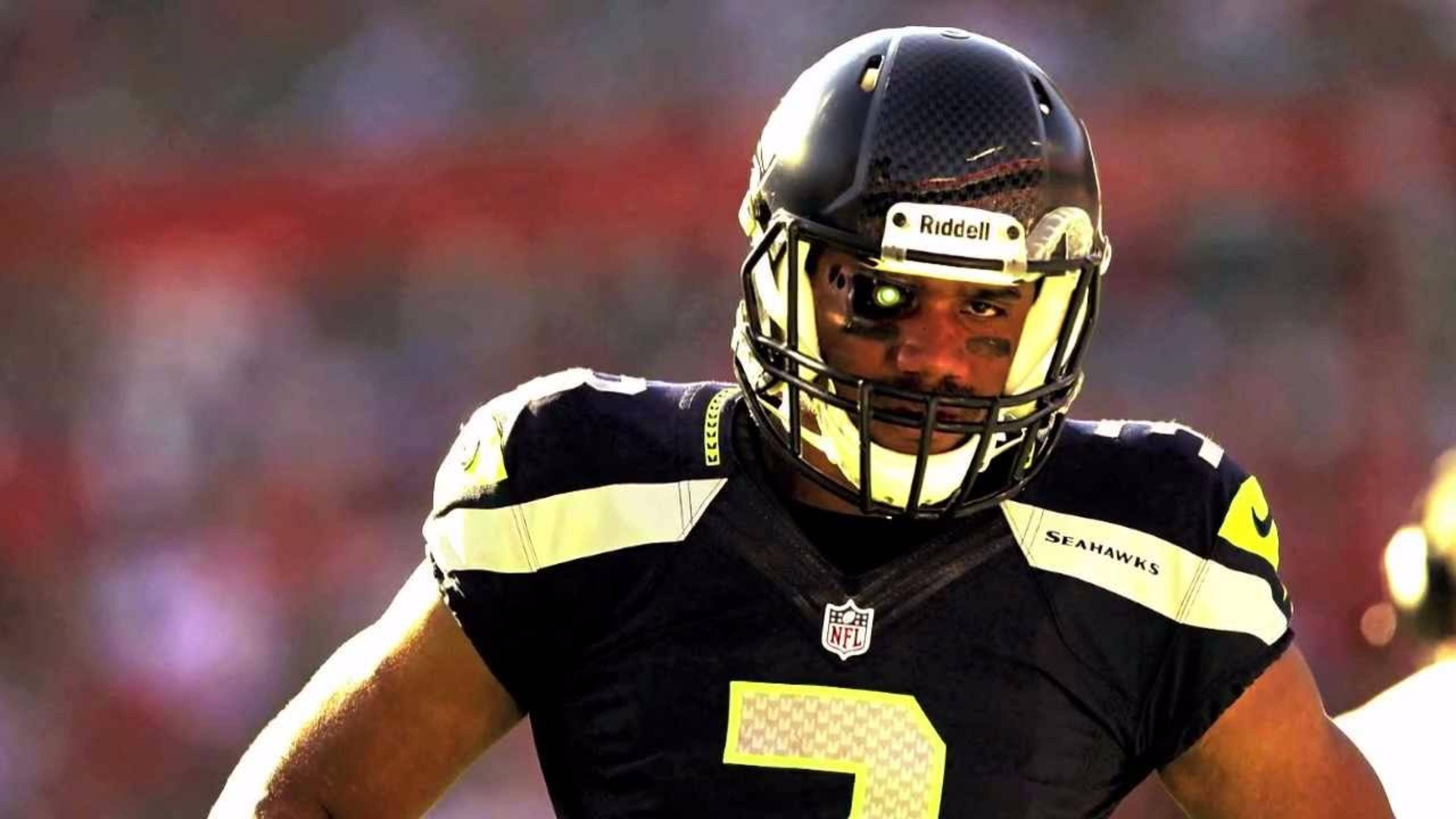 Russell Wilson 嚴肅表情 壁紙 — 一名西雅圖海鷹隊美式足球員身穿黑色和金色球衣，在比賽中透過頭盔凝視著。