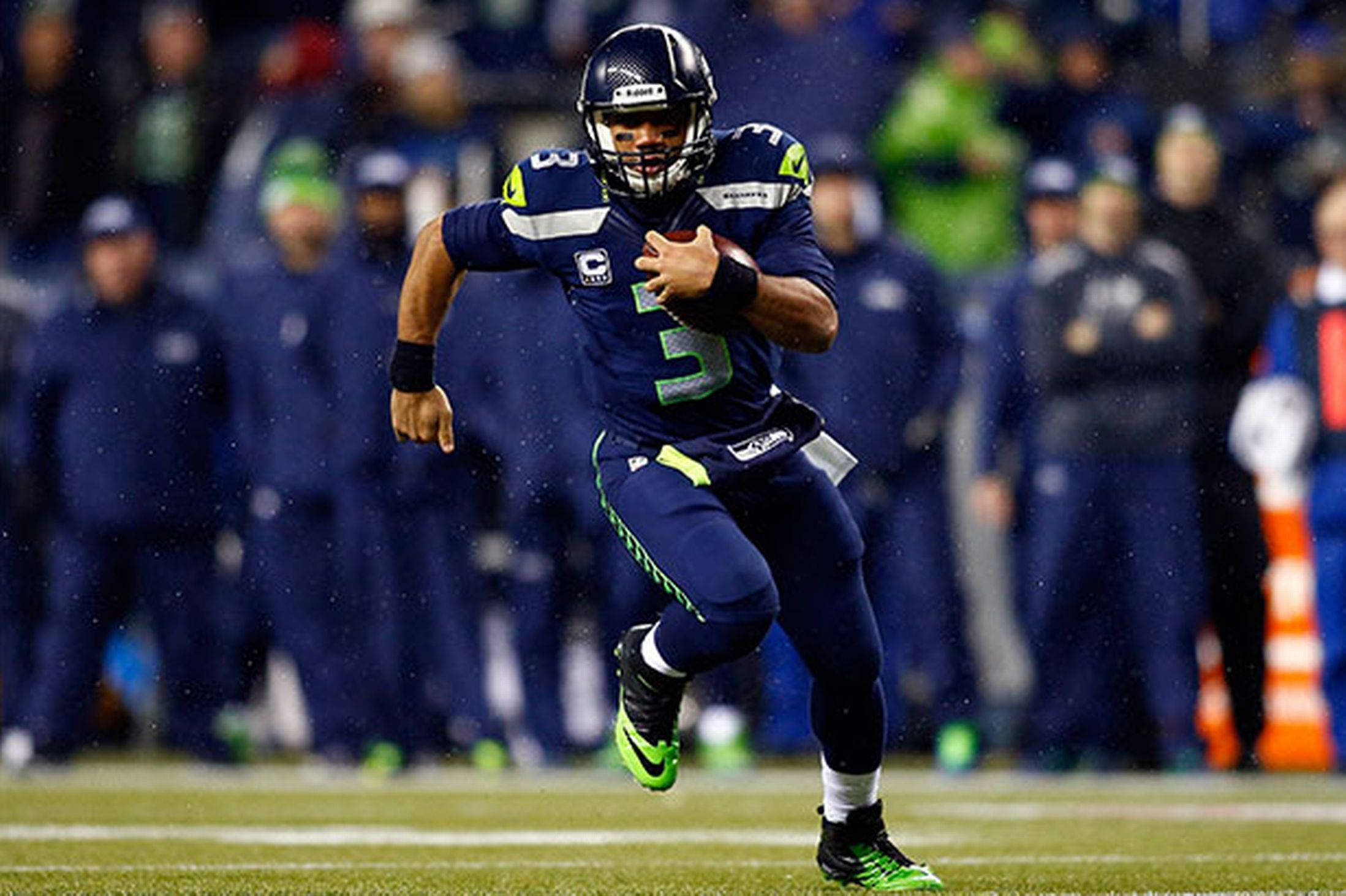 Russell Wilson 雨中奔跑 壁紙 — 西雅圖海鷹隊的四分衛身穿藍綠球衣，在雨夜比賽中持球衝刺。