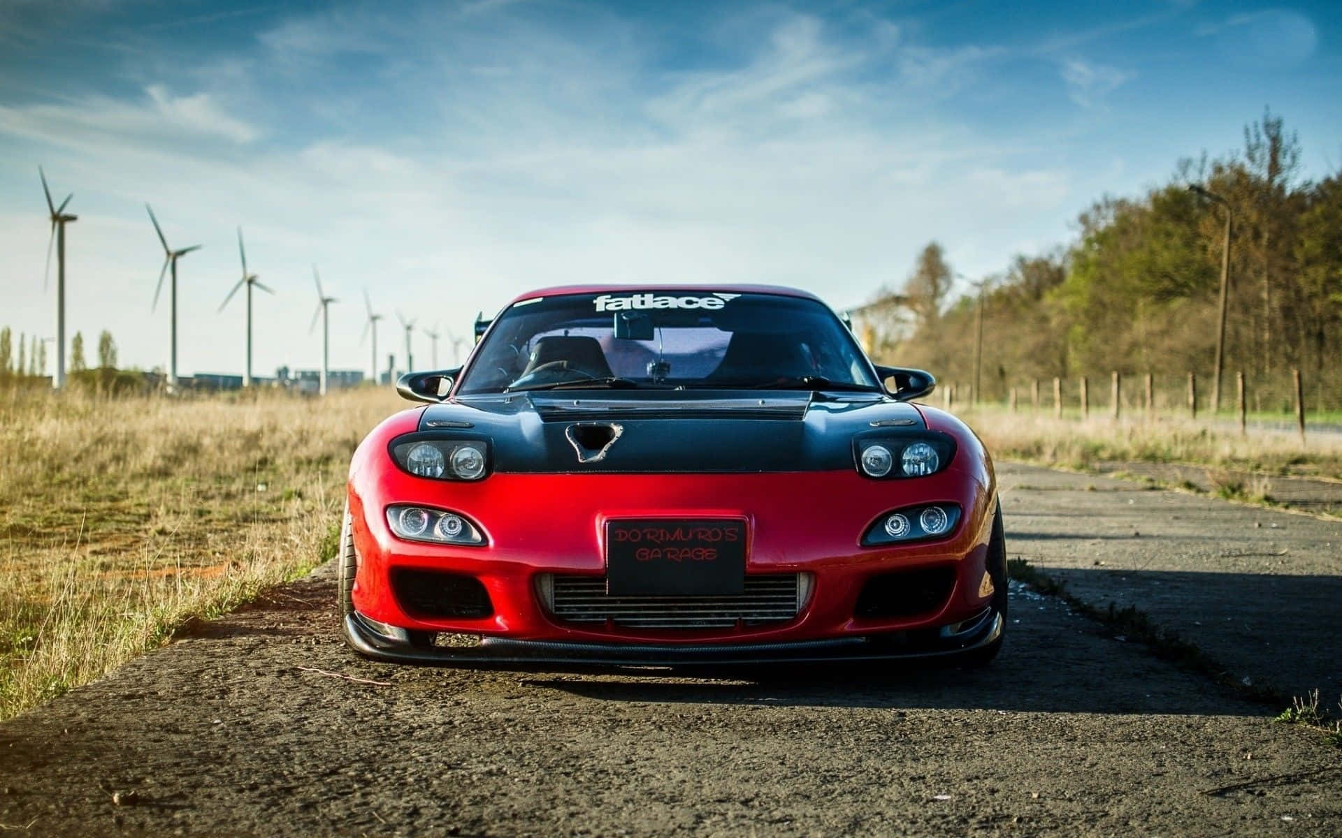 紅色馬自達RX-7對比風車 壁紙 — 一輛紅色和黑色的萬事得RX-7跑車停泊在碎石路上，背景是藍天下的風力發電機。