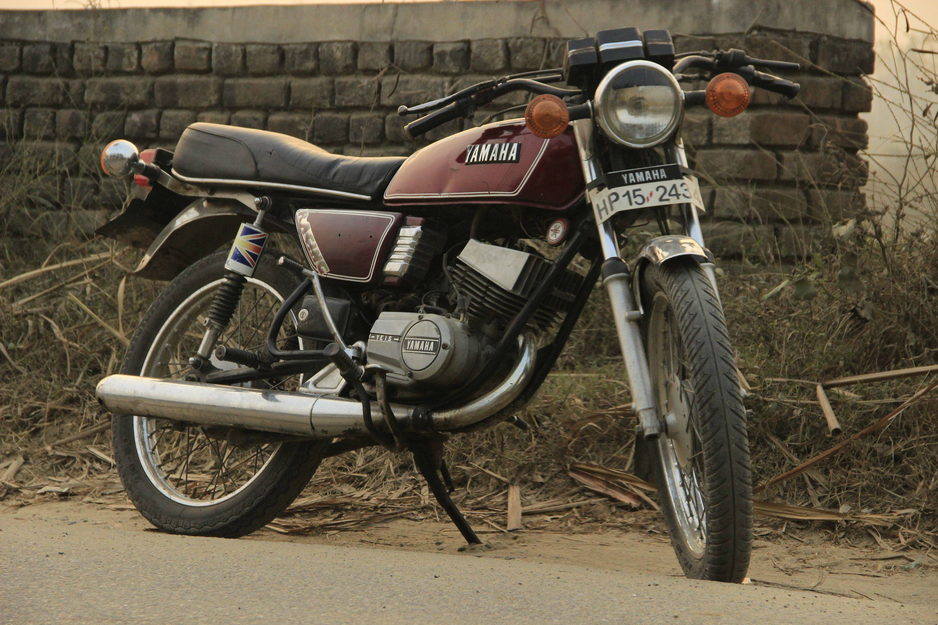 路邊紅色雅馬哈RX100摩托車 壁紙 — 一輛復古酒紅色Yamaha電單車停泊在塵土飛揚的路邊，背景是風化的磚牆。