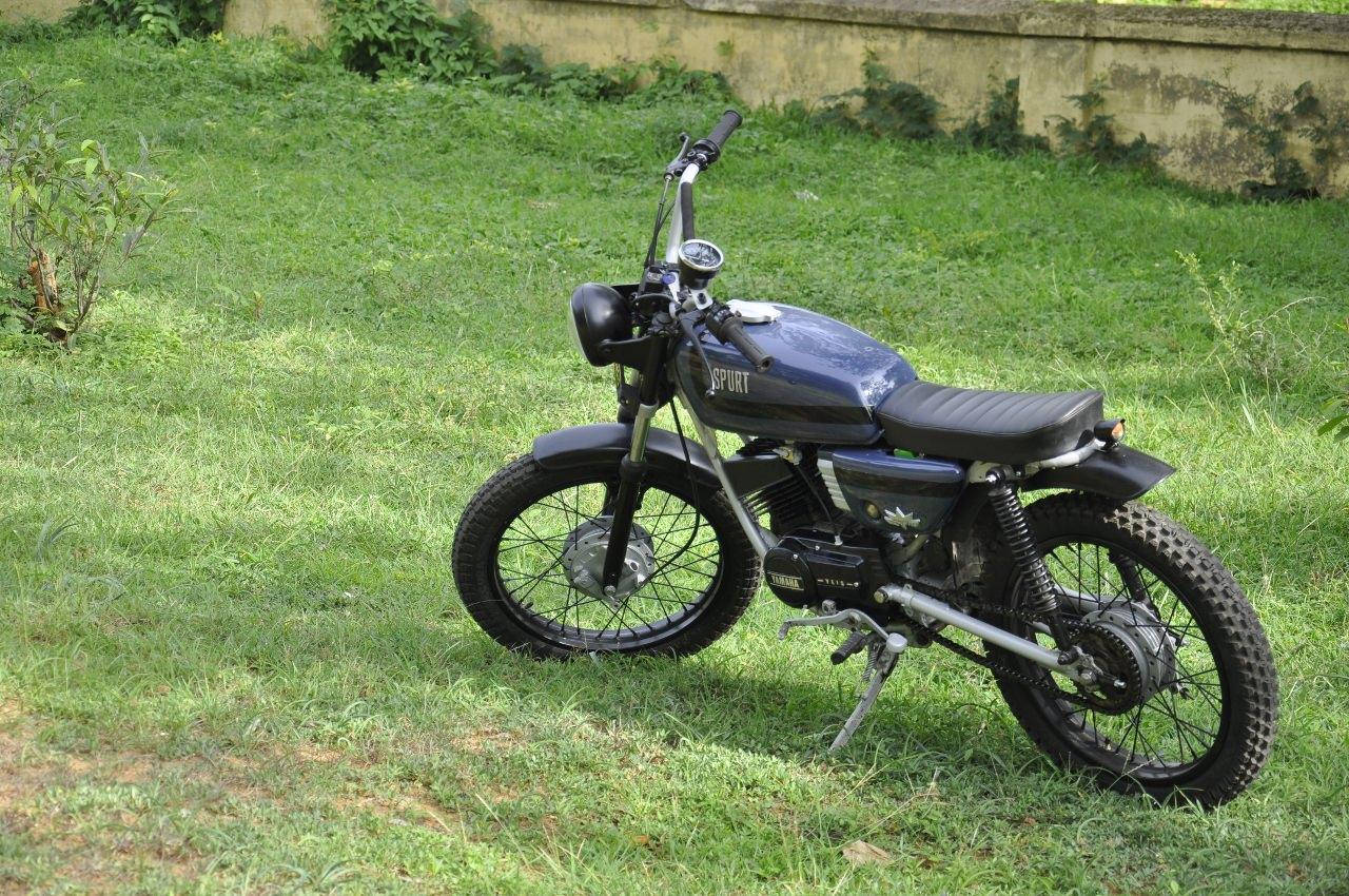 停在庭院中的雅馬哈RX100 Tracker摩托車 壁紙 — 一架時尚黑色復古電單車配備越野輪胎，停泊在陽光普照的草地上，旁邊是風化的混凝土牆。