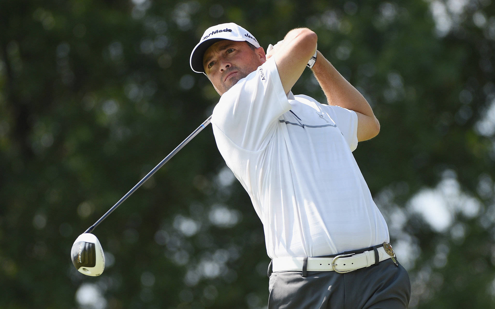 Ryan Palmer 力量揮桿 壁紙 — 一位專注的高爾夫球手在明亮的日光下，以模糊的綠色樹木為背景揮桿。