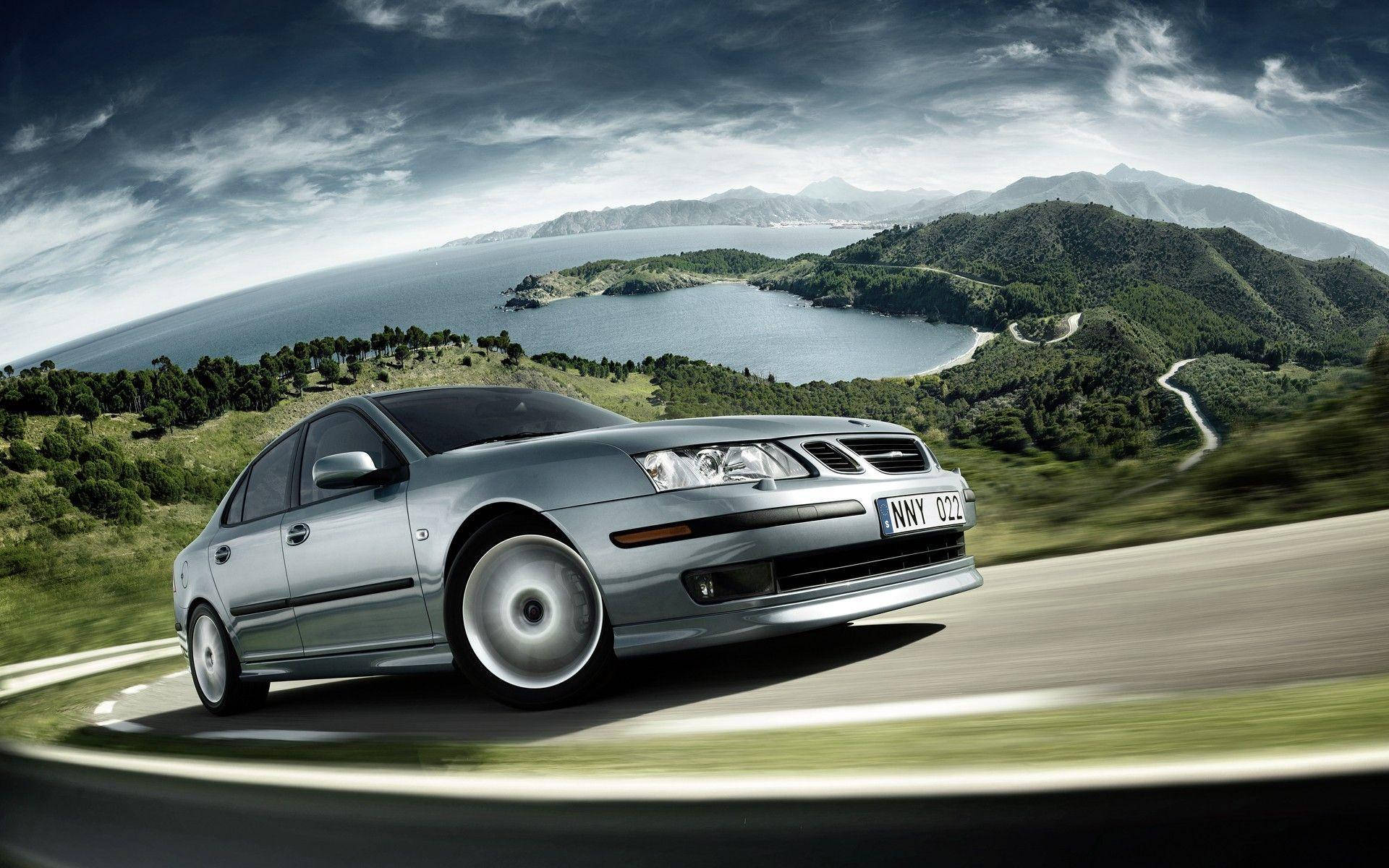 Saab 9-3 Aero Sedan 壁紙 — 一輛銀色房車沿著蜿蜒的海岸山路飛馳，下方是海灣，天空多雲而富有戲劇性。