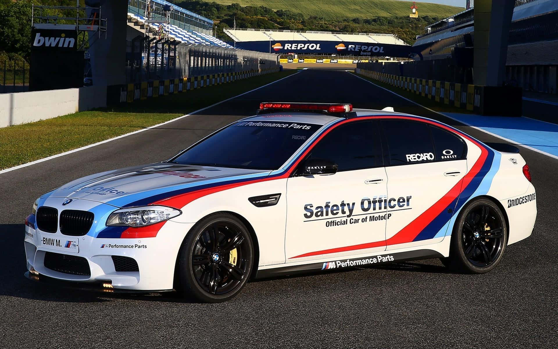 賽道上亮眼的Safety Car 壁紙 — 一輛帶有紅藍條紋的白色BMW M5安全車在陽光明媚的賽道上奔馳，賽道兩旁有看台和Repsol品牌標誌。