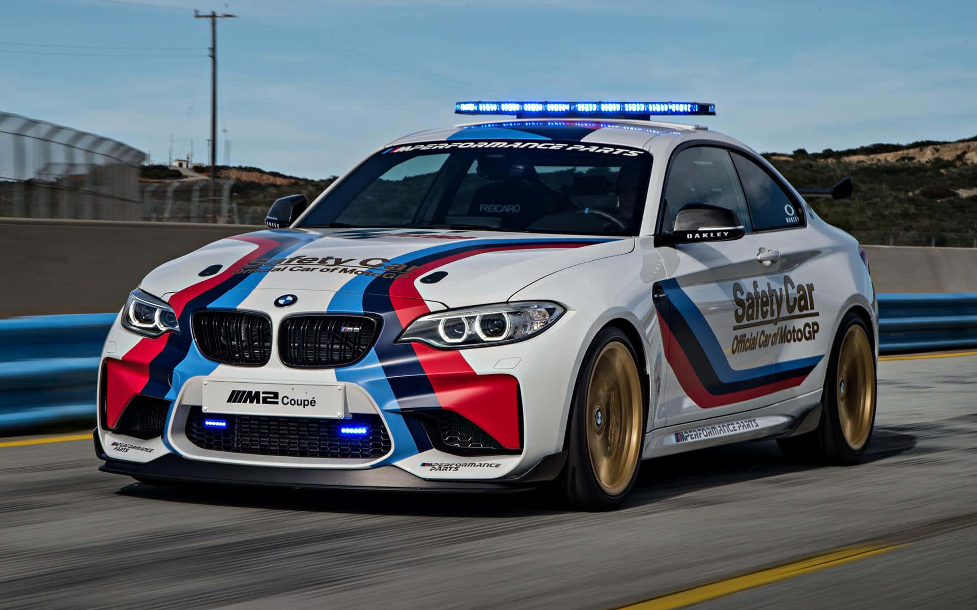 速度與安全——賽道上優雅的Safety Car 壁紙 — 一輛帶有藍色、紅色和黑色賽車條紋的白色BMW M2 Coupe安全車，在晴朗的藍天下沿著賽道飛馳。